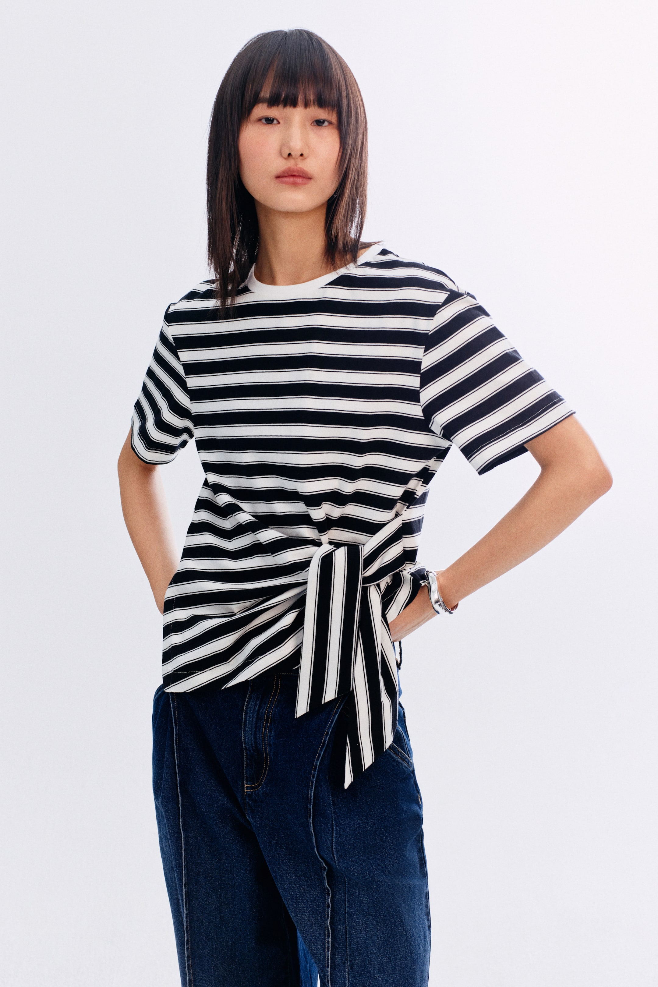 拡大画像を表示: 黒と白のストライプ柄のTシャツは、ショートスリーブとラウンドネックラインを特徴としており、左腰でフロントを結んで、ギャザーの入った非対称のディテールを作り出しています。