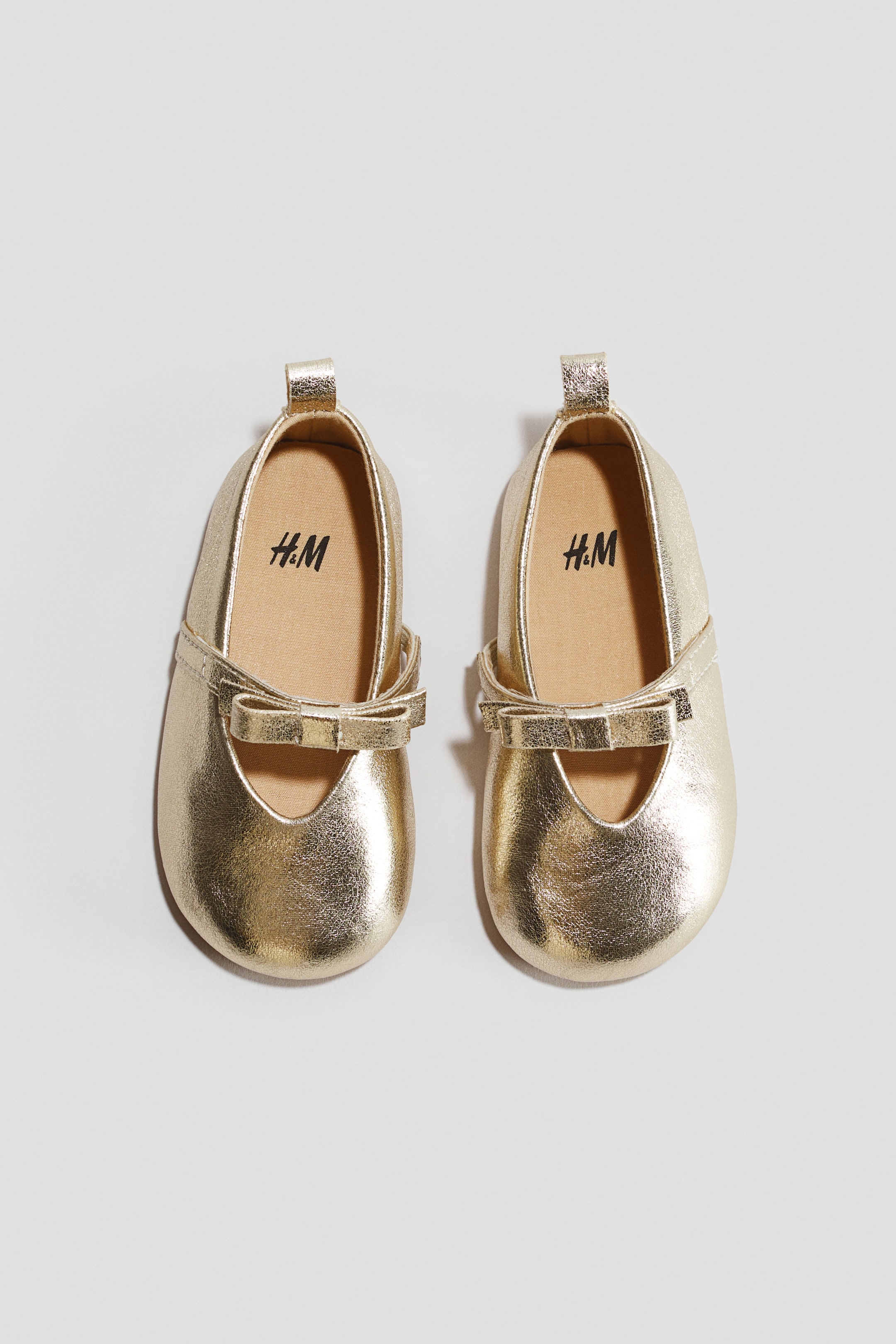 Voir une image plus grande: Une paire de ballerines dorées est orientée vers l'avant, avec des bouts arrondis et une lanière ornée d'un nœud sur le pied. Une petite boucle est visible à l'arrière de chaque talon, et les semelles intérieures brun clair affichent le logo H&M.