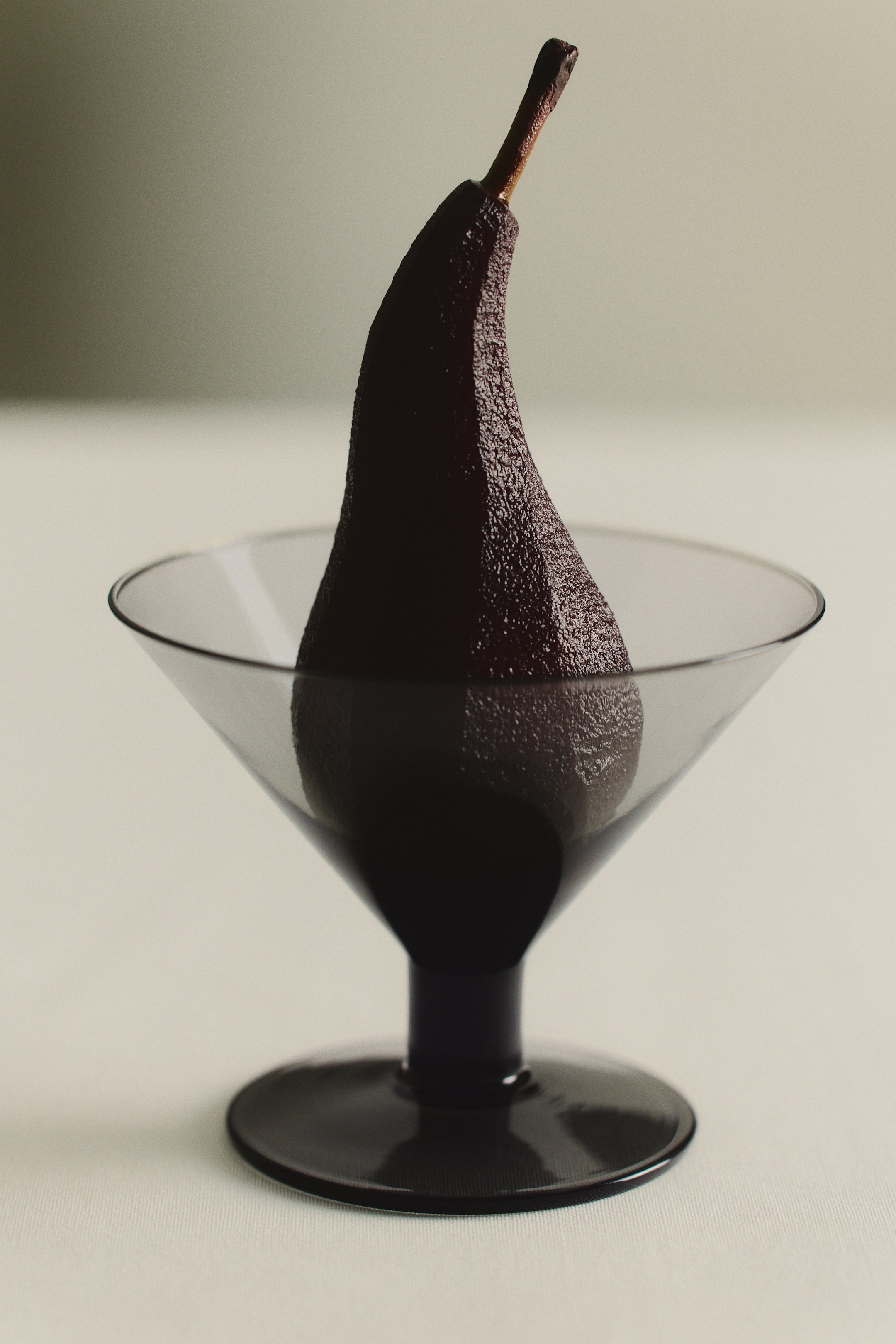 Ver imagen más grande: Se muestra de frente una copa de cóctel soplada a boca, con cáliz cónico, un tallo corto de tono oscuro y una base circular plana, que contiene en su interior una pera pochada oscura, colocada en posición vertical.