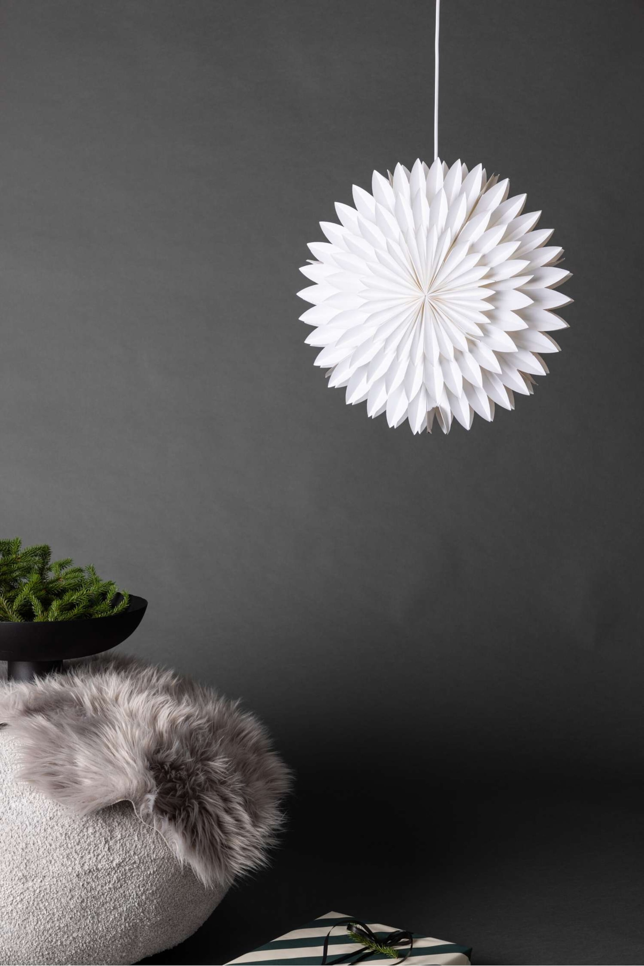 Größeres Bild anzeigen: Eine kugelförmige weiße Papier-Hängedekoration mit schichtweise angeordneten, spitz zulaufenden Falten, die sich in einem blumenähnlichen Muster nach außen entfalten, hängt an einer dünnen weißen Schnur.