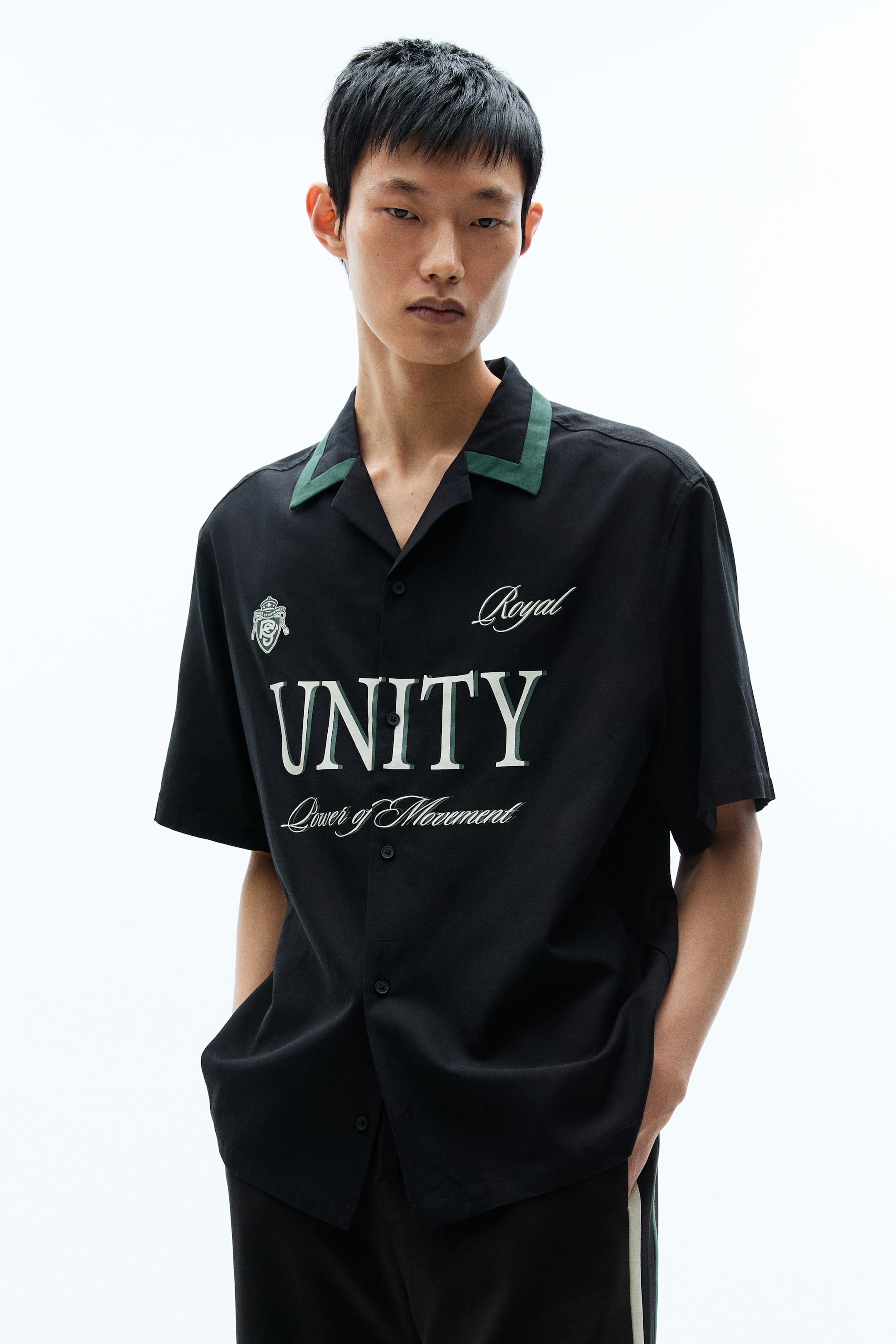 View larger image: Front view of a black short-sleeved shirt, featuring a green-trimmed camp collar and a full button-front placket. The chest displays "Royal" in script, an emblem, and "UNITY Power of Movement" in white and green lettering.