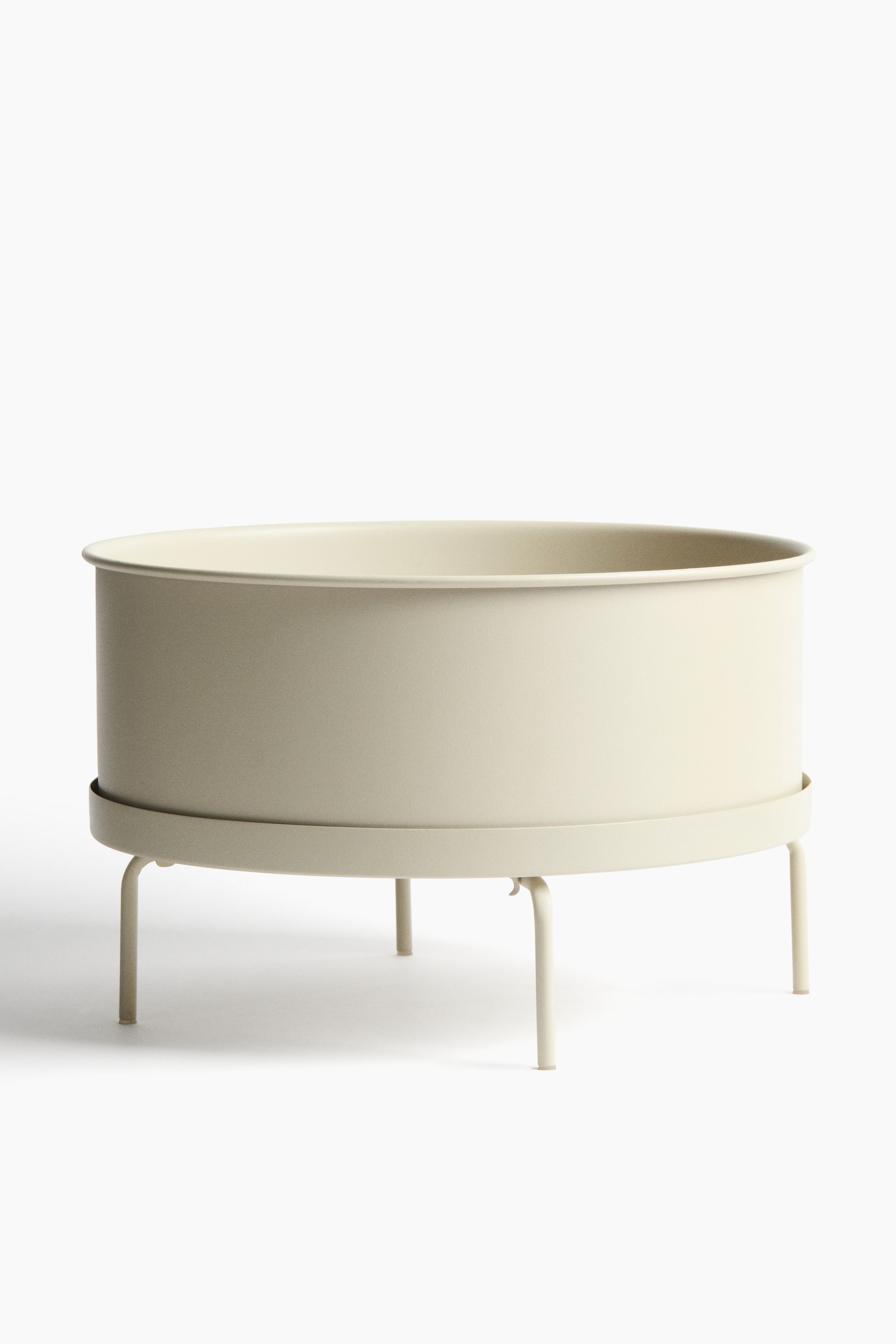 View larger image: Front view of a cylindrical, light-beige metal plant pot seated in a matching metal plant stand. The stand has a saucer supporting the pot and four curved legs.