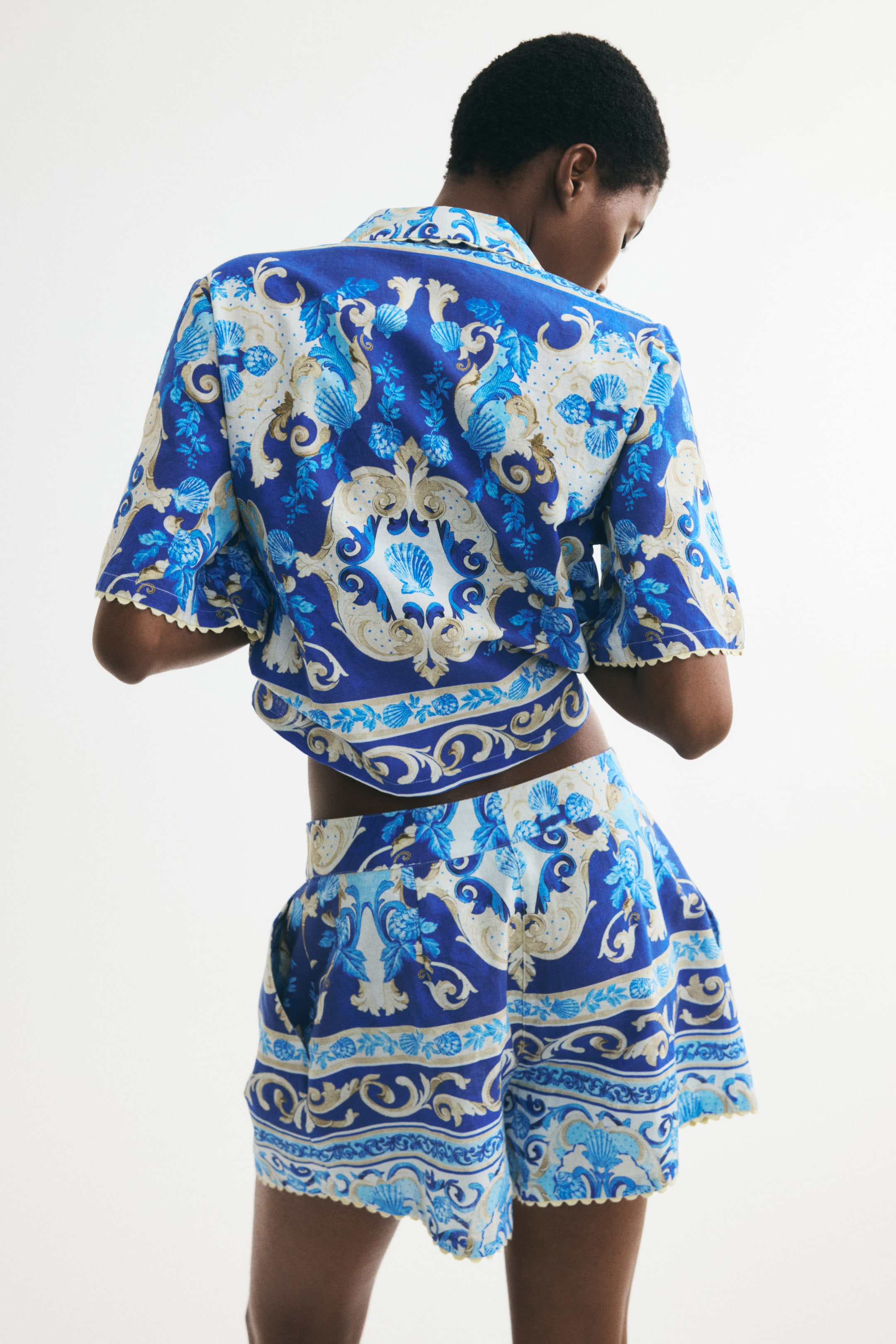 Ver imagem maior: Um conjunto de duas peças, composto por uma blusa de manga curta e calções, é apresentado de costas, exibindo um padrão em estilo barroco de azul, branco e dourado com conchas. A blusa cropped apresenta uma gola e bordas recortadas nas mangas e na bainha, enquanto os calções mostram uma cintura lisa, bolsos laterais e bainhas recortadas.