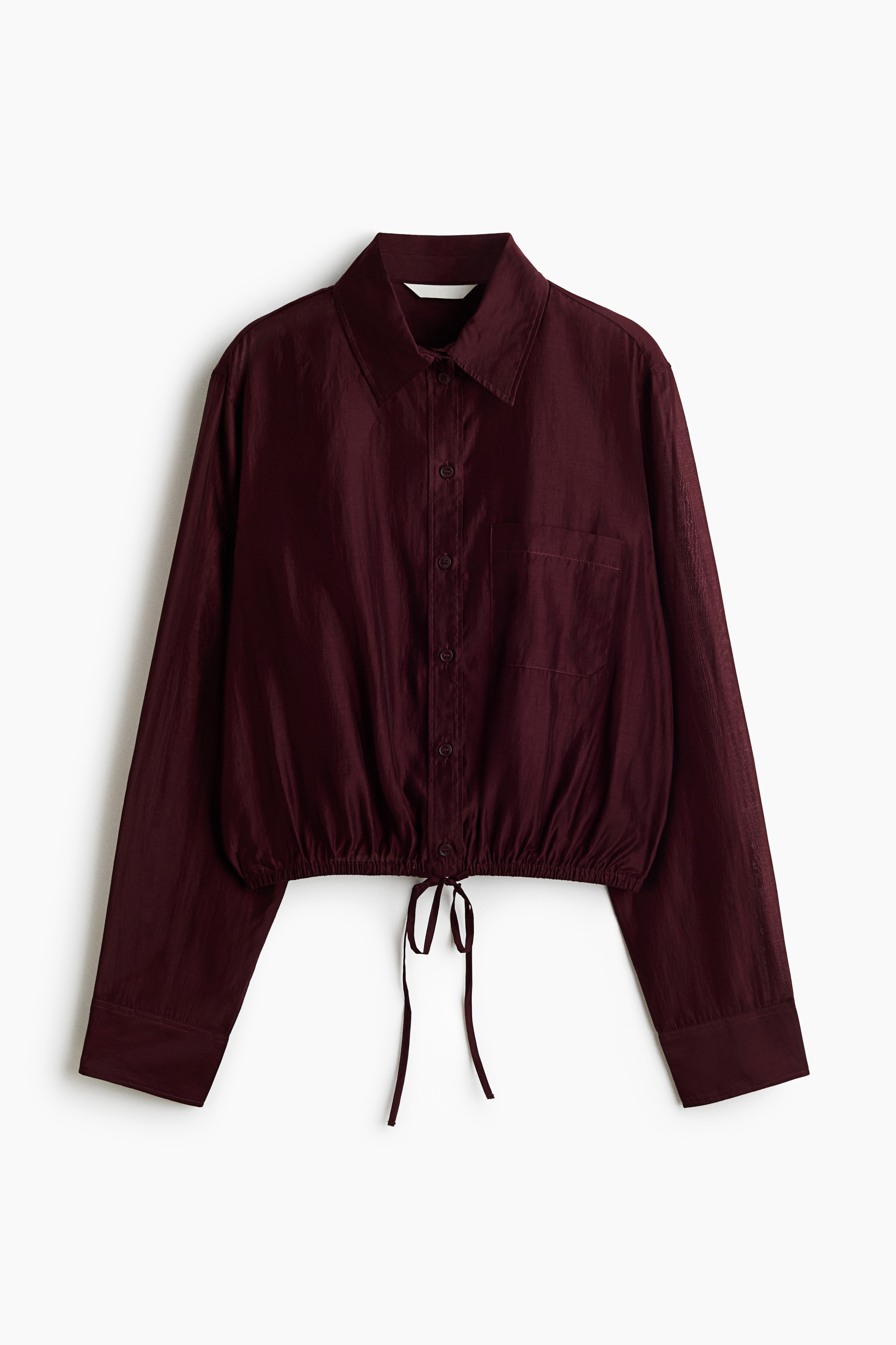 Một chiếc áo sơ mi crop màu đỏ burgundy với độ bóng nhẹ đang được người mẫu mặc, có cổ áo, cúc cài phía trước, một túi patch ở ngực và gấu áo bo thun với dây rút buộc ở eo. Áo được phối cùng quần jeans đen ống rộng.
