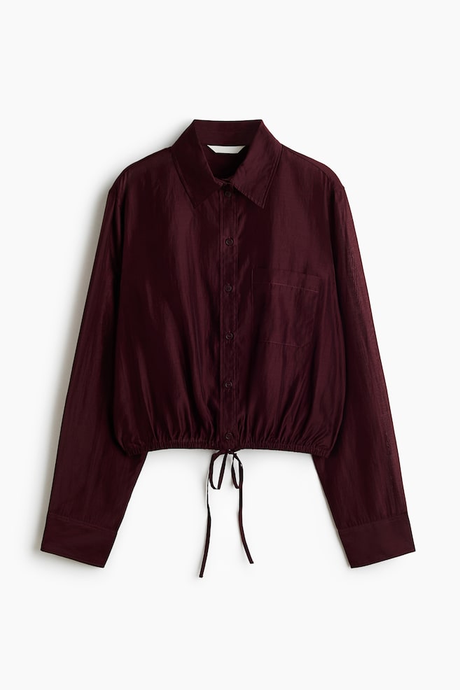 A burgundy cropped button-down shirt is shown from the front, featuring a point collar, a single chest patch pocket, and long sleeves with wide cuffs. The shirt is gathering at the hem with an elasticized band and a matching thin drawstring tie at the center. - 1