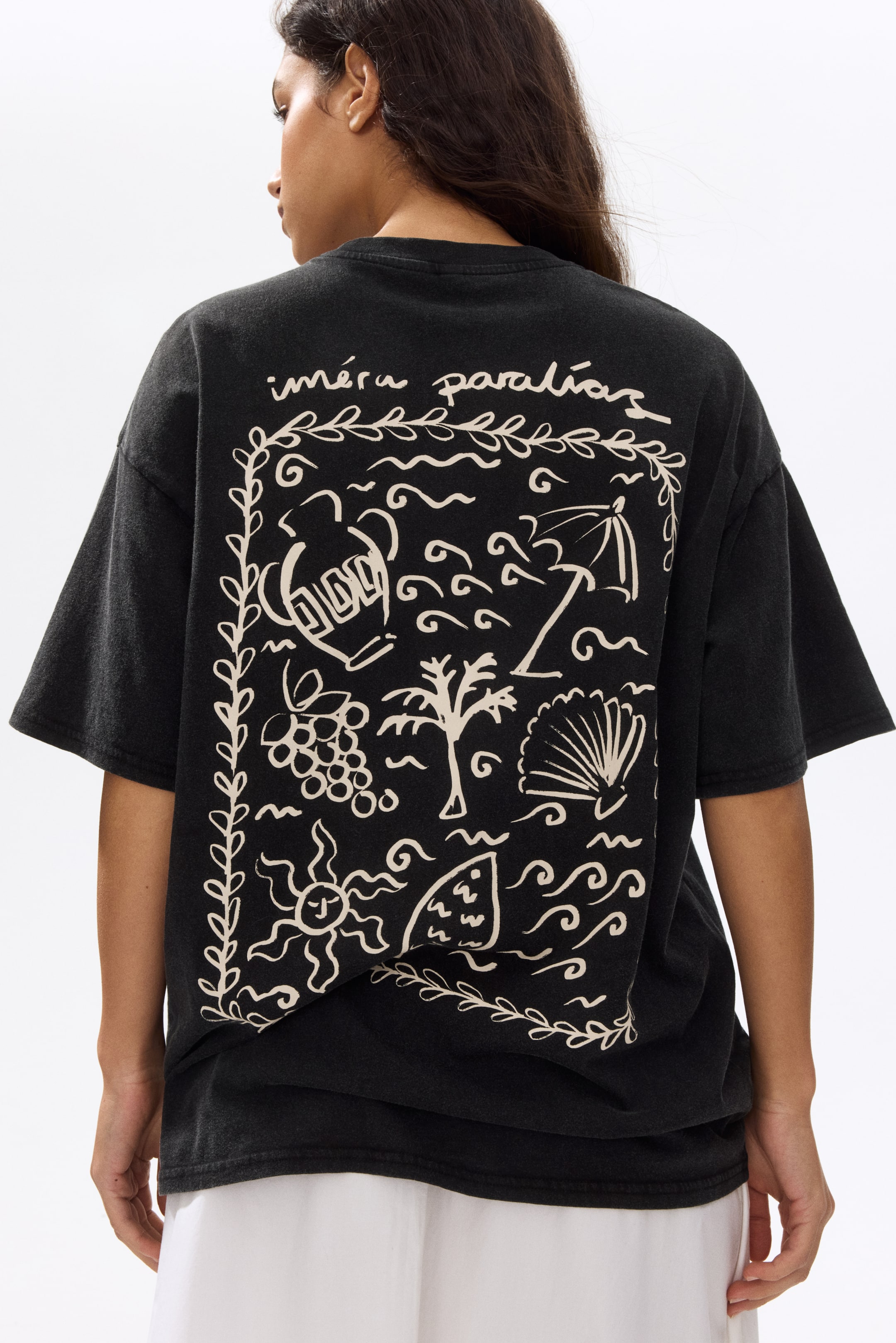 View larger image: The model is wearing an oversized black cotton jersey t-shirt, showcasing dropped shoulders, a straight hem, and a prominent, light-coloured print motif on the back. The print features abstract and nature-inspired elements within a leafy border, with text placed above. The t-shirt is styled over a white garment.