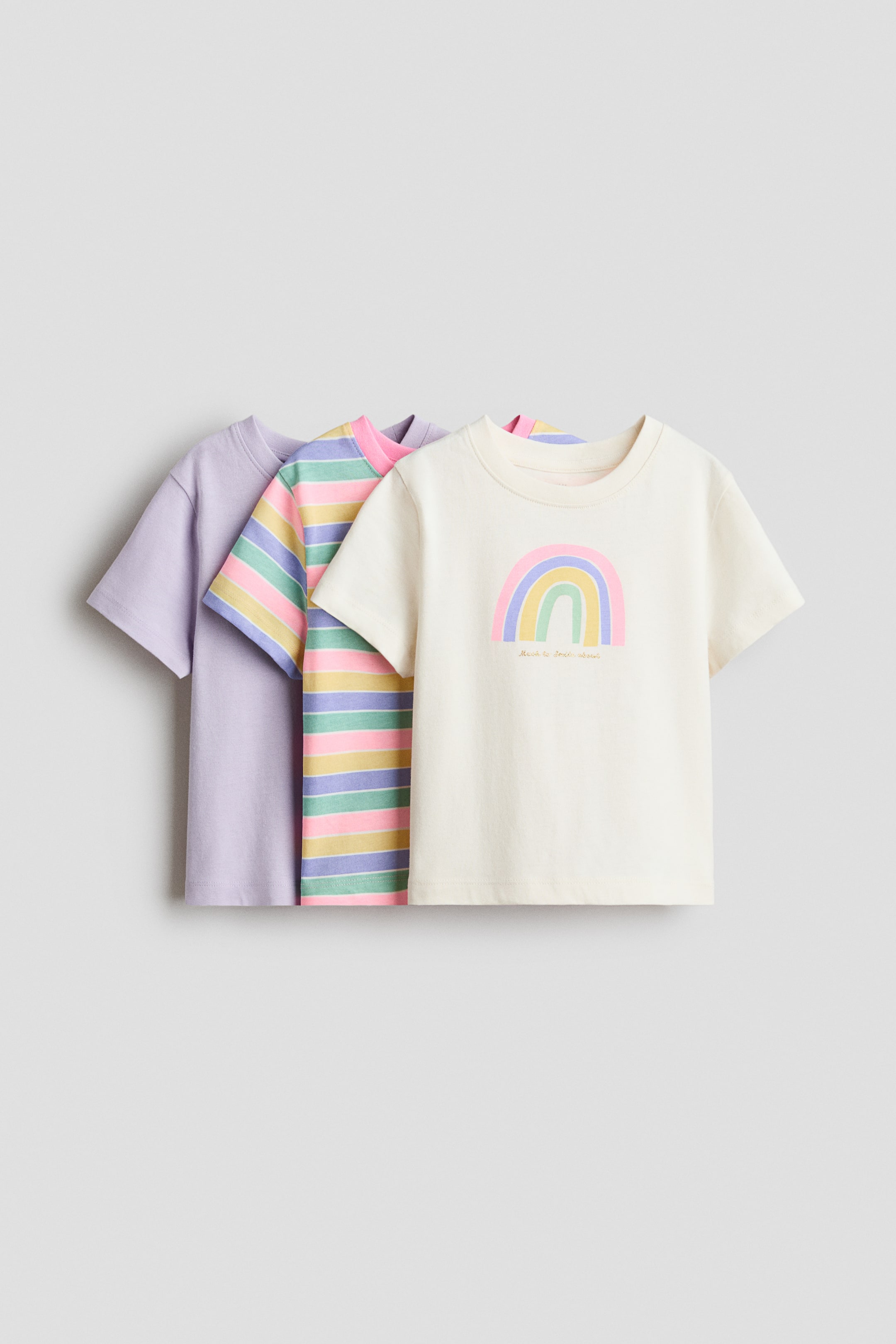 View larger image: "T-shirts crafted from soft cotton jersey, featuring a round, ribbed neckline, are presented as a set of three with their fronts facing forward. One t-shirt is in a solid light purple shade, another showcases horizontal stripes in pink, light blue, yellow, and light green, while the third is an off-white shirt adorned with a pastel rainbow graphic on the chest."
