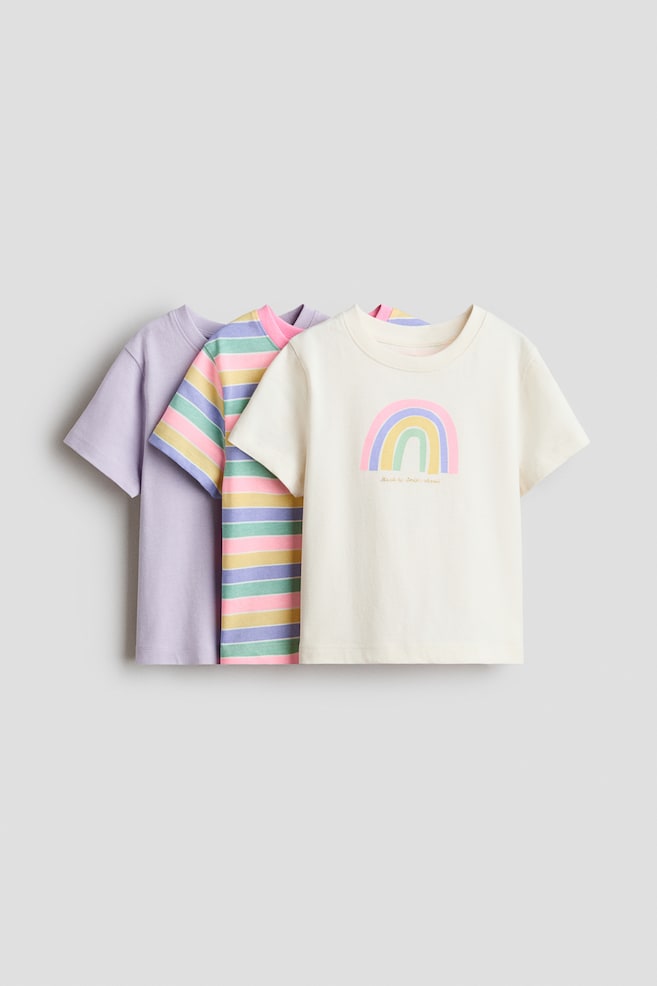 Camisetas de suave jersey de algodón con un cuello redondo ribeteado se presentan en un conjunto de tres, con sus frentes visibles. Una camiseta es de un color lila sólido, otra presenta rayas horizontales en rosa, azul claro, amarillo y verde claro, y la tercera es una camiseta blanco hueso con un gráfico de arcoíris en tonos pastel en el pecho. - 1