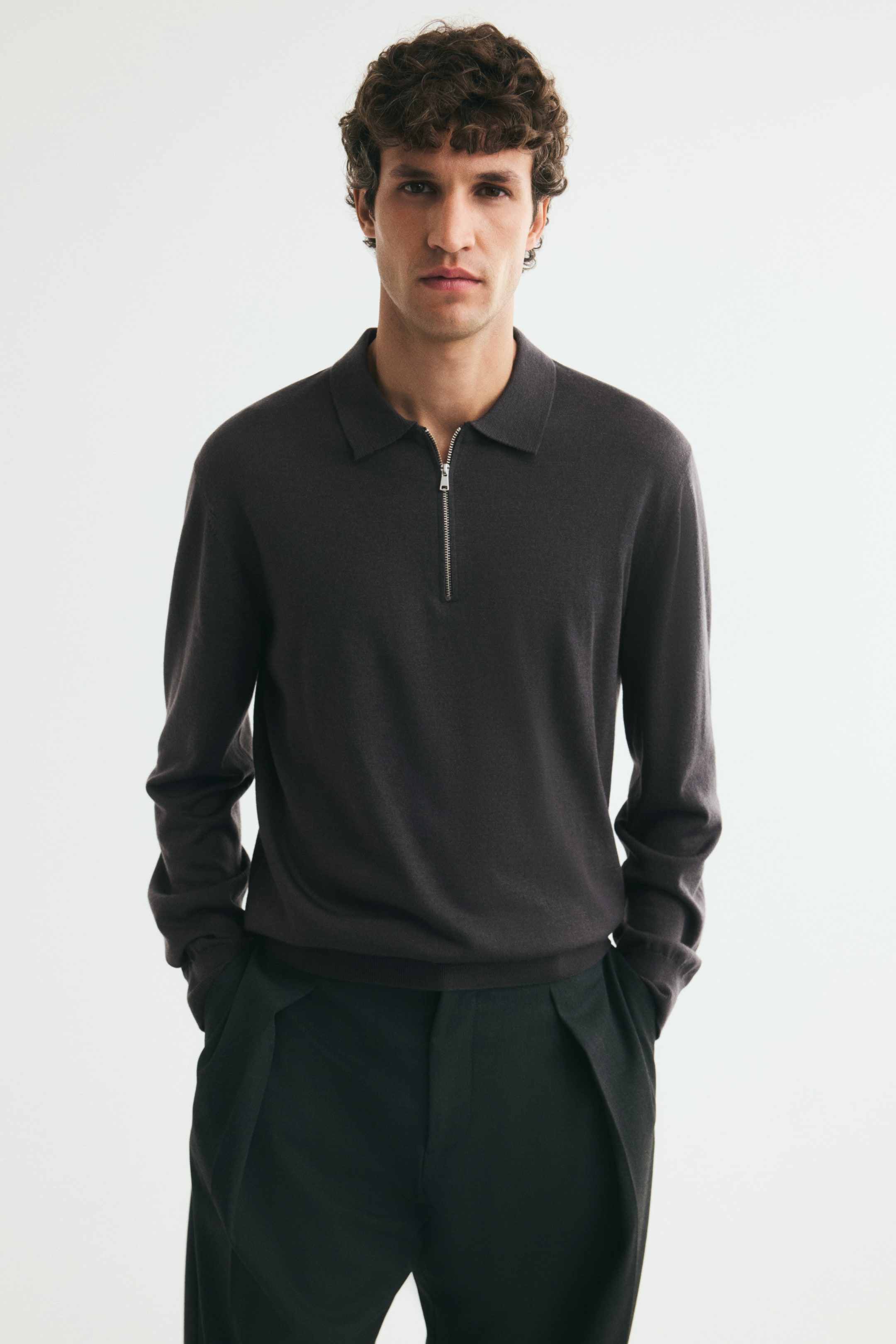 View larger image: A dark grey, fine-knit long-sleeved polo shirt is being worn by the model, who is standing with hands in the pockets of dark trousers. The shirt features a classic collar with a partially open silver half-zip at the neckline and ribbed cuffs.