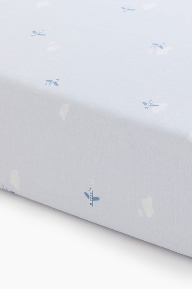 A fitted cot sheet in light blue cotton is shown covering a mattress corner from an angled view, highlighting its snug fit. The fabric features an all-over pattern of small, scattered blue airplanes and white clouds. - 1