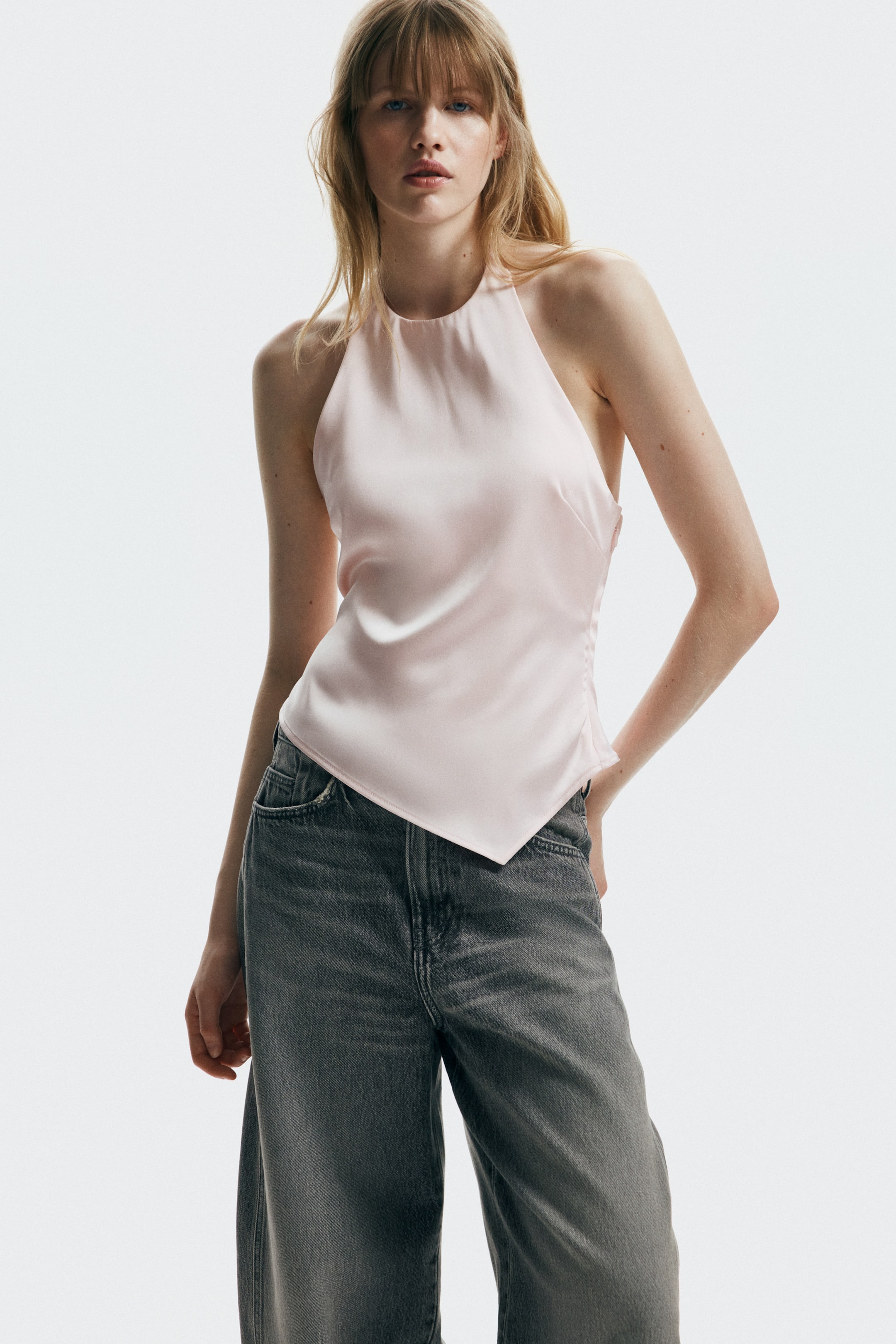 Größeres Bild anzeigen: Ein hellrosa Satin-Top mit Neckholder wird präsentiert, das über einen hohen, abgerundeten Ausschnitt verfügt. Der asymmetrische Saum bildet eine spitze Form an der vorderen linken Seite, während dezente Raffungen die linke Seite zieren und eine drapierte Textur erzeugen.