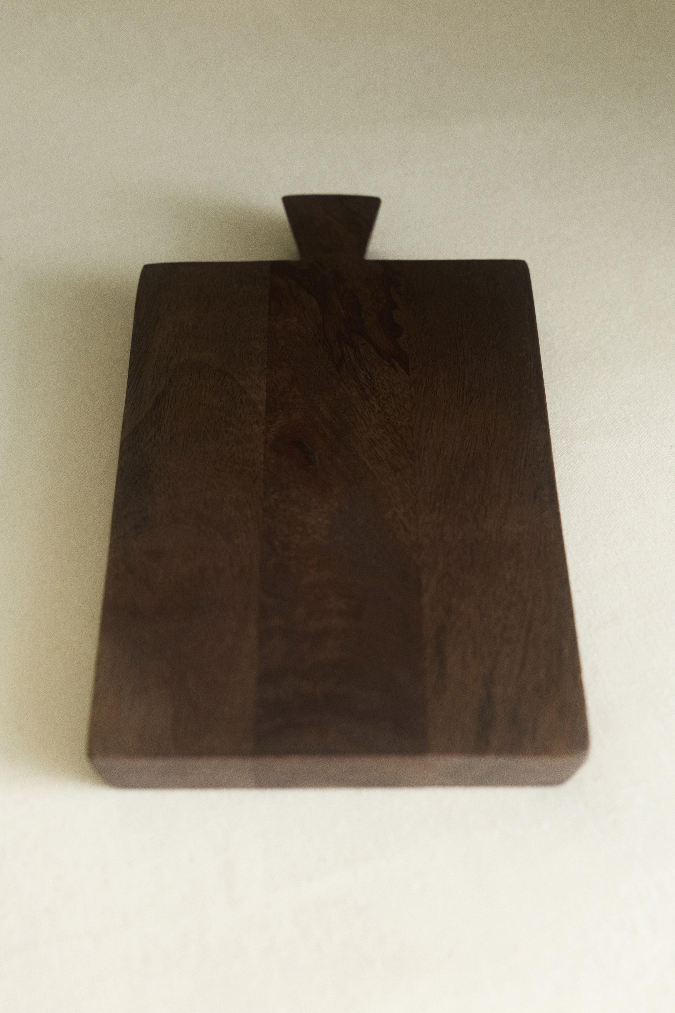 View larger image: A rectangular serving board made from dark mango wood is shown from the front, showcasing its natural wood grain and smooth surface. The board features a short, flared handle at the top centre.