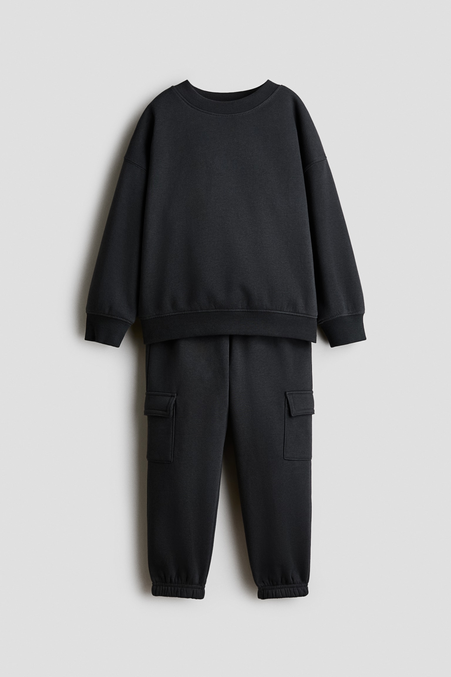 Ett svart tvådelat träningsset visas framifrån och består av en långärmad sweatshirt och matchande sweatpants. Sweatshirten har en ribbad rund halsringning, nedhasade axlar samt ribbade muddar och fåll, medan sweatpantsen har fickor med lock på sidorna och elastiska benslut vid anklarna.