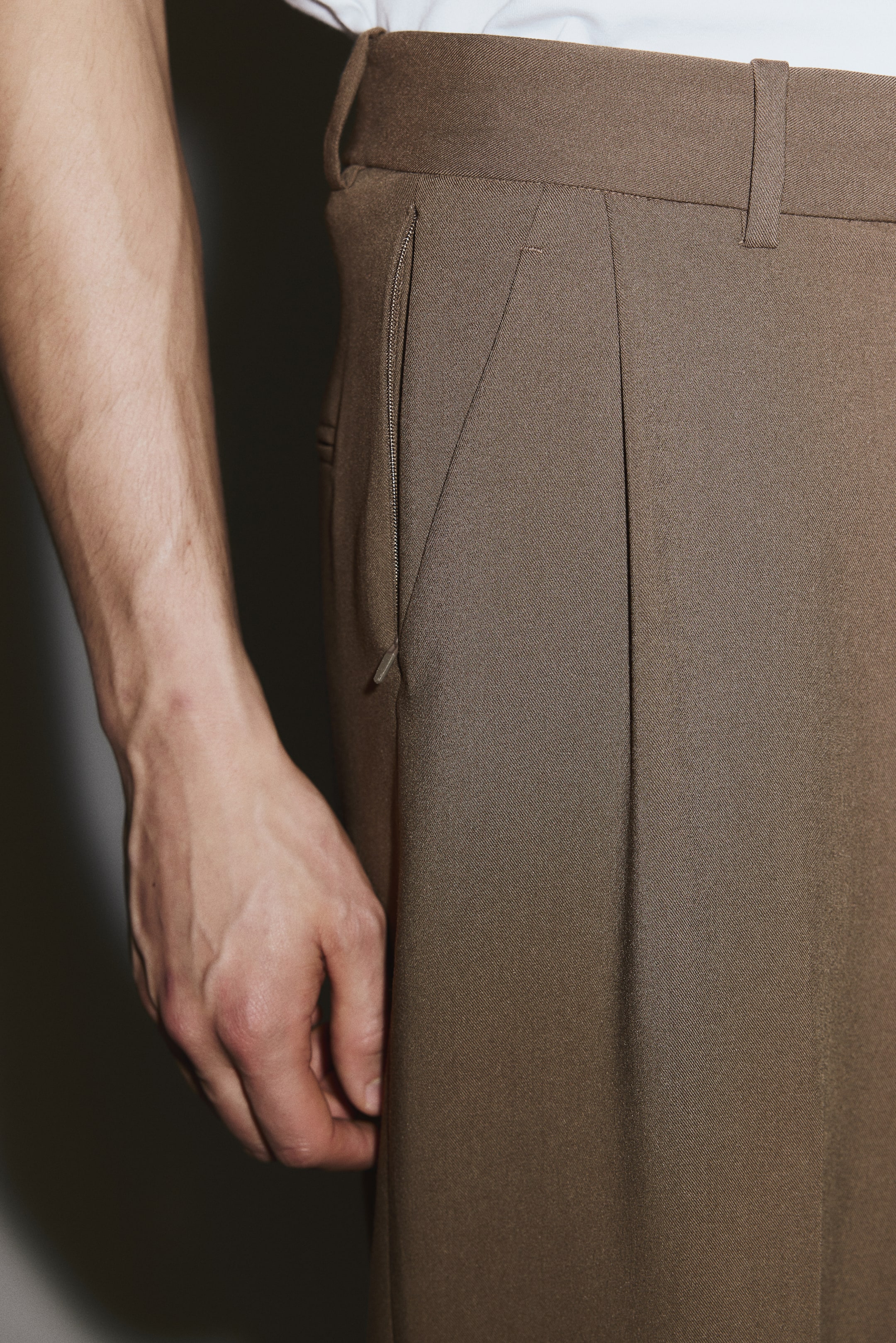 View larger image: Brown trousers are shown from a side angle, with a flat waistband and belt loops, and a white shirt tucked into them. A zippered side-seam pocket is visible, and a single front pleat runs vertically down the leg.