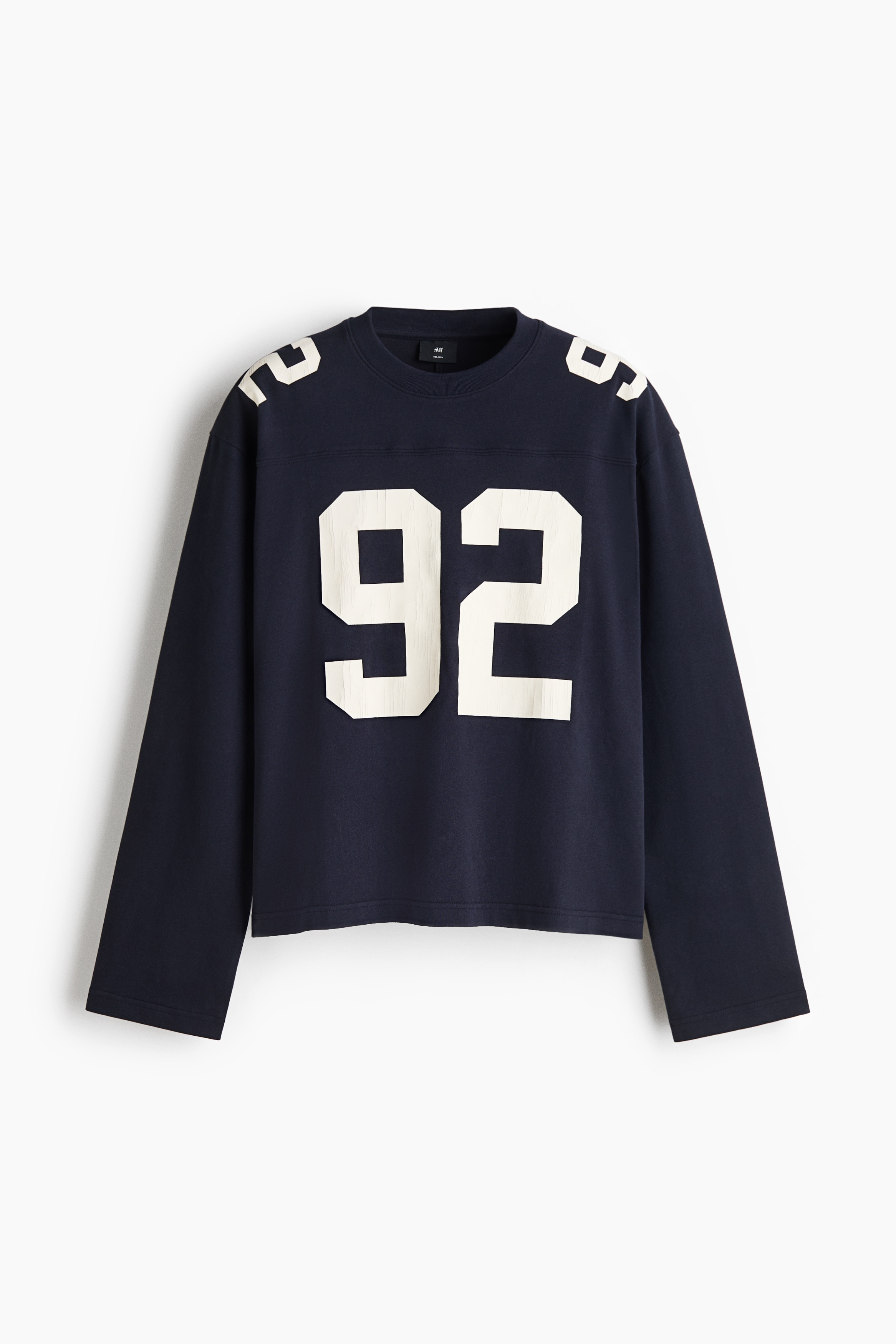 A dark blue long-sleeve tee in a relaxed fit with a front yoke is being worn by the model. The tee features a large white number "92" printed on the chest, matching white prints on the shoulders, and a ribbed round neckline layered over a white collar.