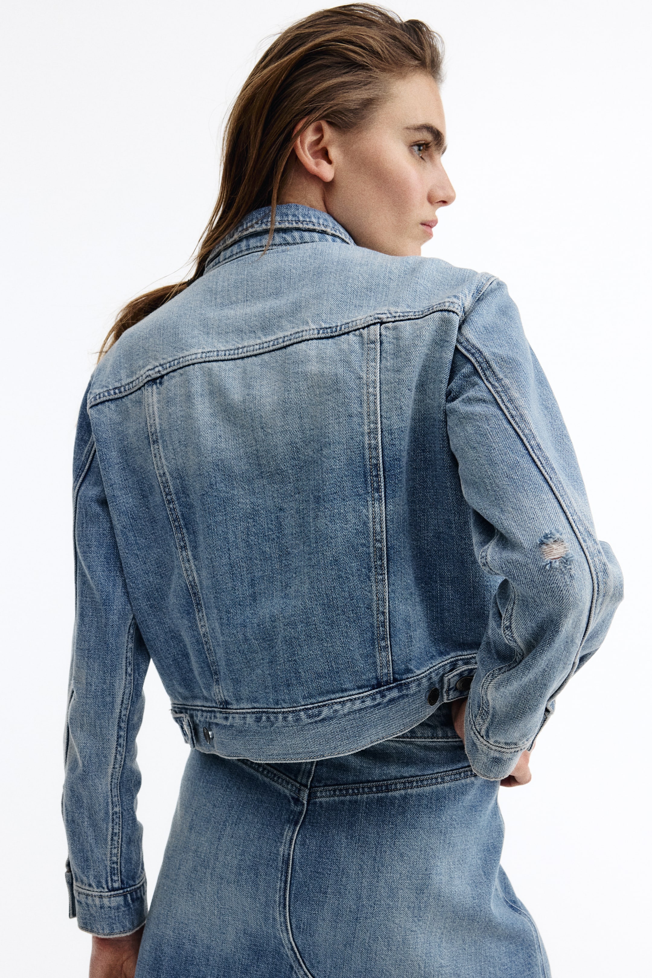 View larger image: The light blue denim jacket is angled slightly to the right from the back view, showcasing a pointed collar, a horizontal back yoke with vertical seams, and adjustable button tabs on the bottom hem. The left sleeve features a distressed detail near the elbow.