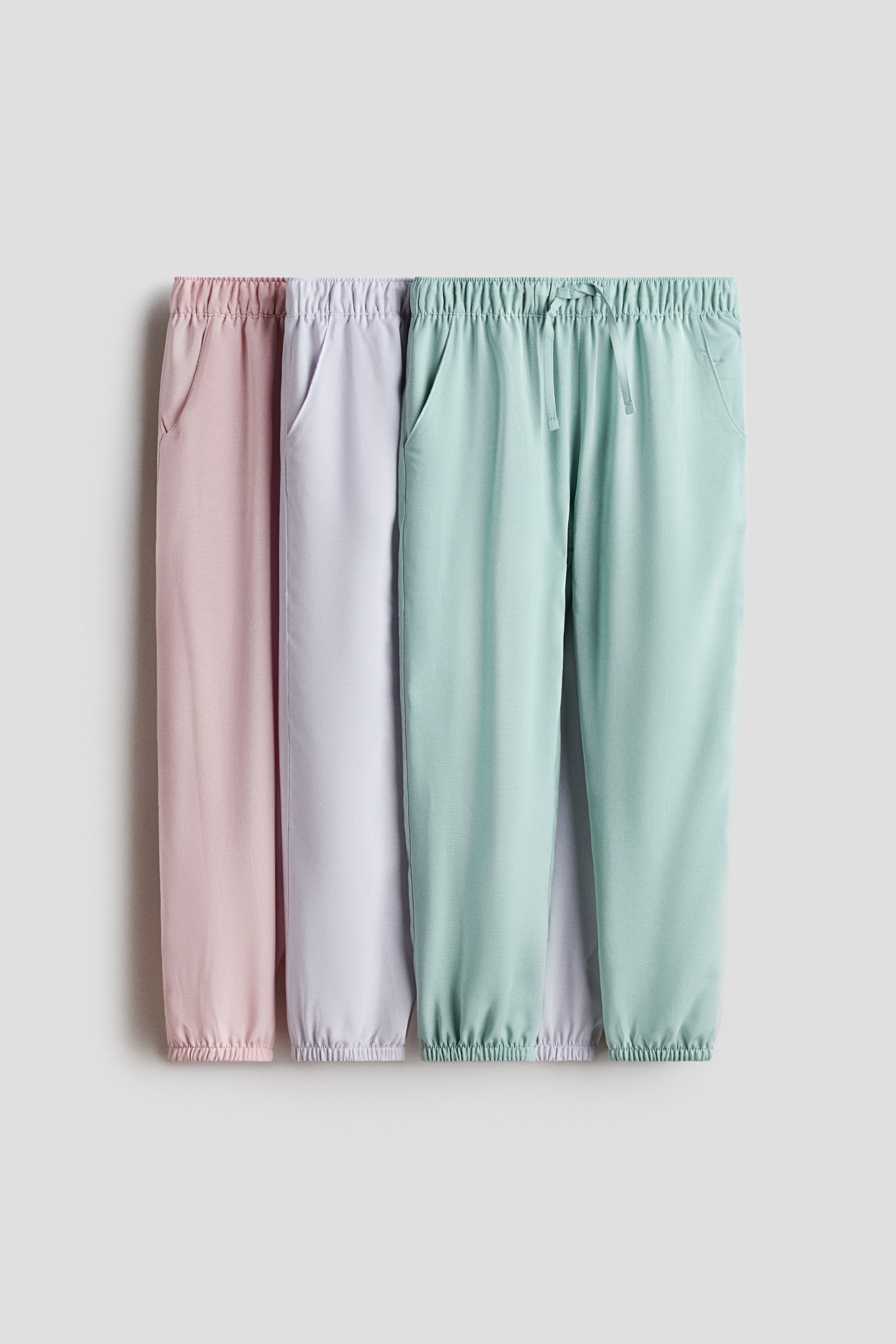 Agrandir l'image: Trois paires de joggeurs en tissage de viscose léger sont suspendues, vues de face. Elles présentent une taille élastiquée avec un nœud décoratif, des poches latérales, des jambes fuselées et une élastication recouverte aux ourlets, et sont proposées en rose, violet clair et vert menthe.