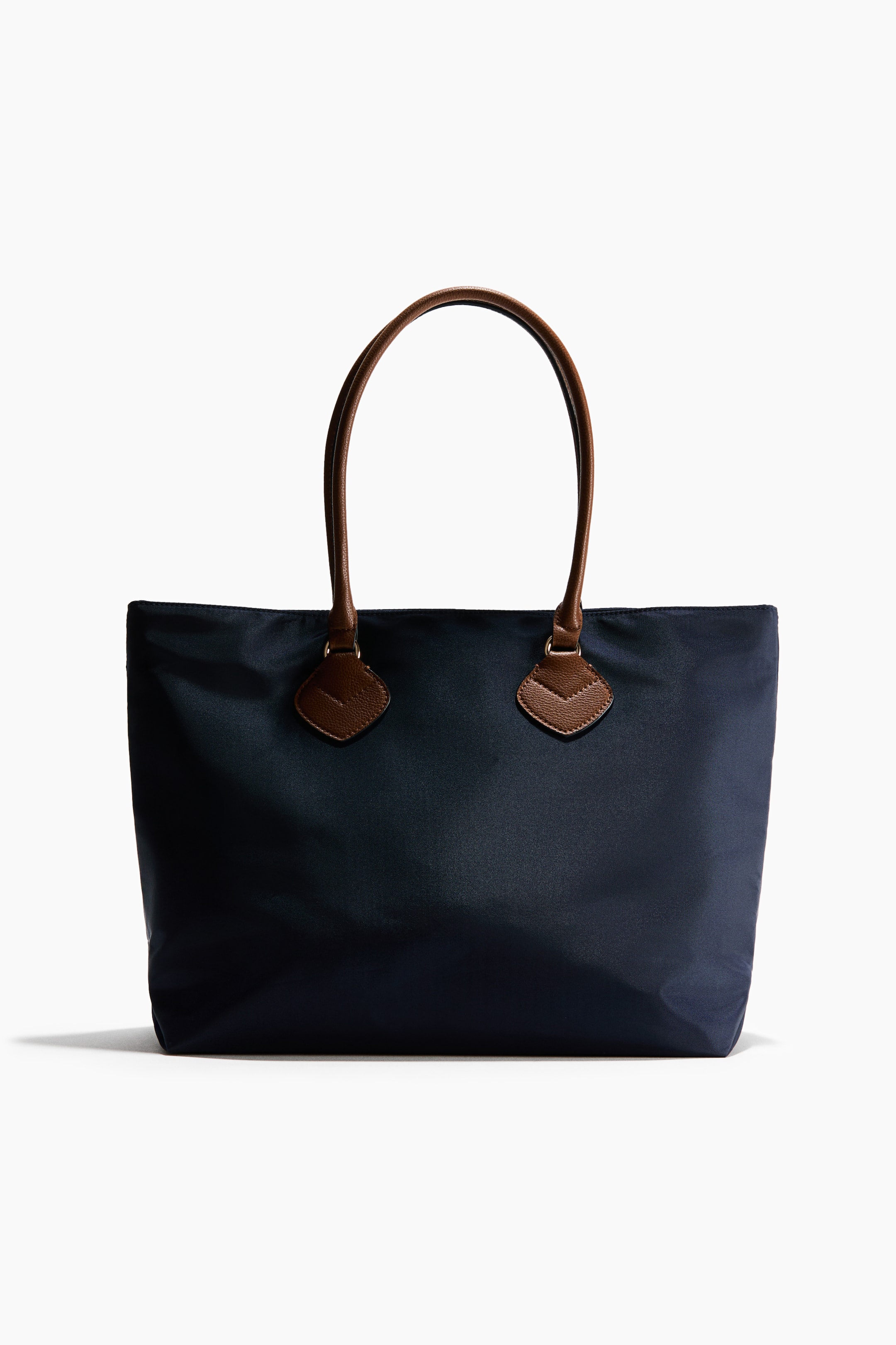 View larger image: A navy blue fabric tote bag is shown facing forward, highlighting its rectangular shape and smooth finish. The bag features two brown textured leather handles that loop upward, secured to the front with gold-tone hardware and diamond-shaped leather patches.