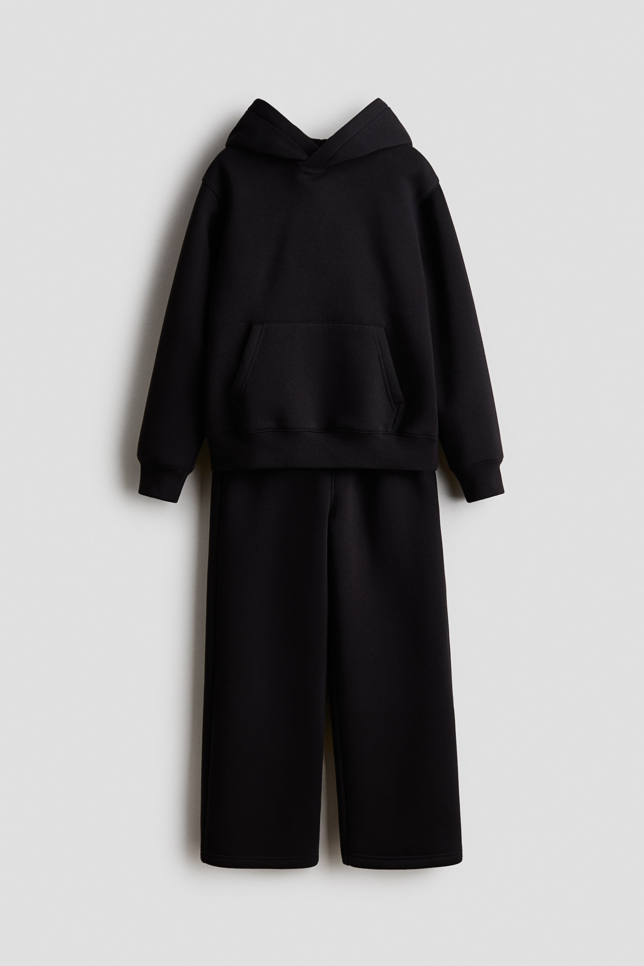 查看大圖: 一套黑色兩件式家居服正面展示,包括一件套頭連帽衫,配有交叉式帽兜、前置袋鼠袋及羅紋袖口。配套直筒褲整齊地掛在連帽衫下方,完整呈現整體搭配套裝。