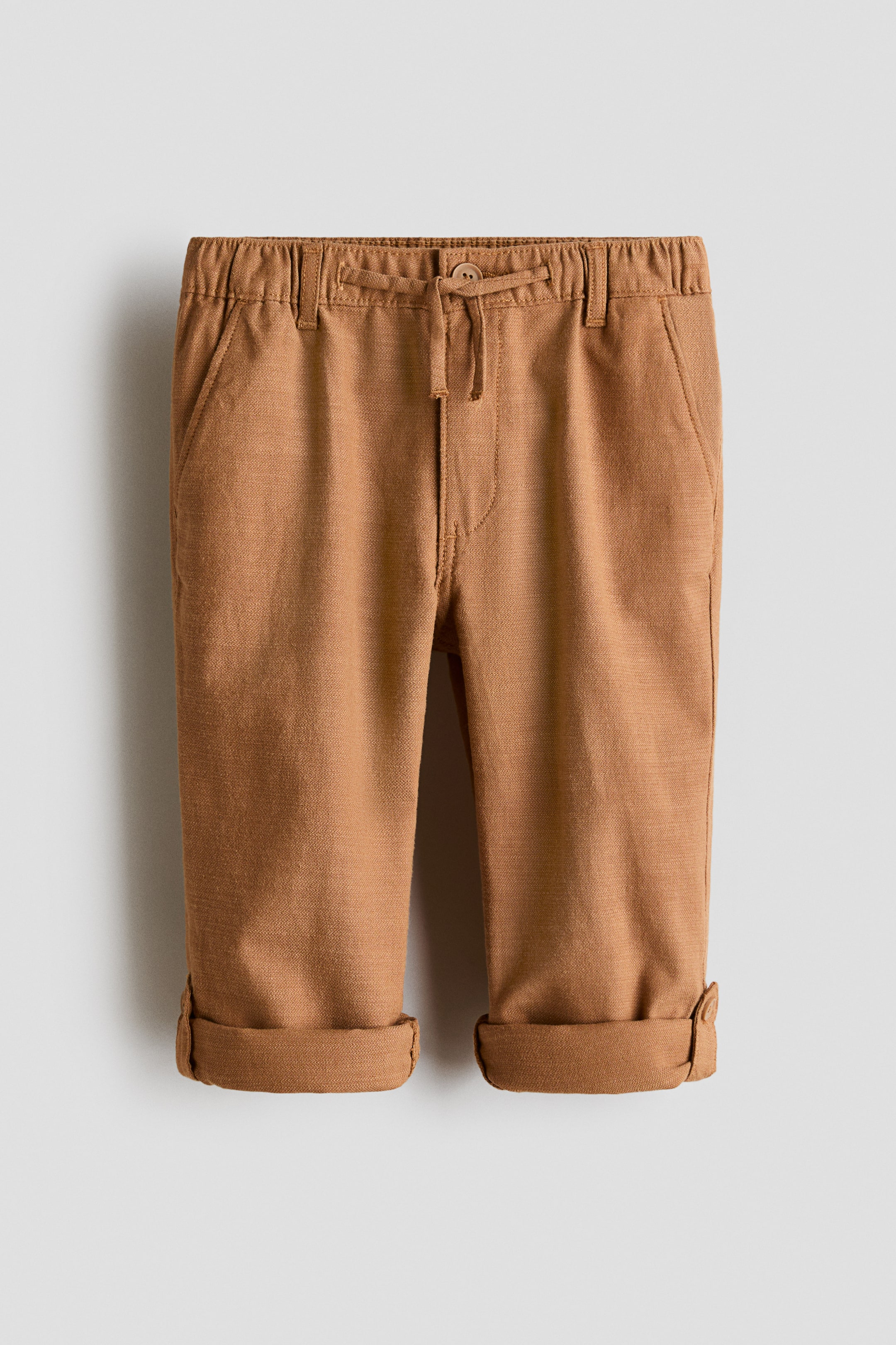 View larger image: Trousers in textured cotton twill are shown from the front, highlighting a loose fit through the hip and thigh with tapered legs. They feature an elastic drawstring waist with a faux fly, diagonal side pockets, and rolled-up cuffs with a button tab on one side.