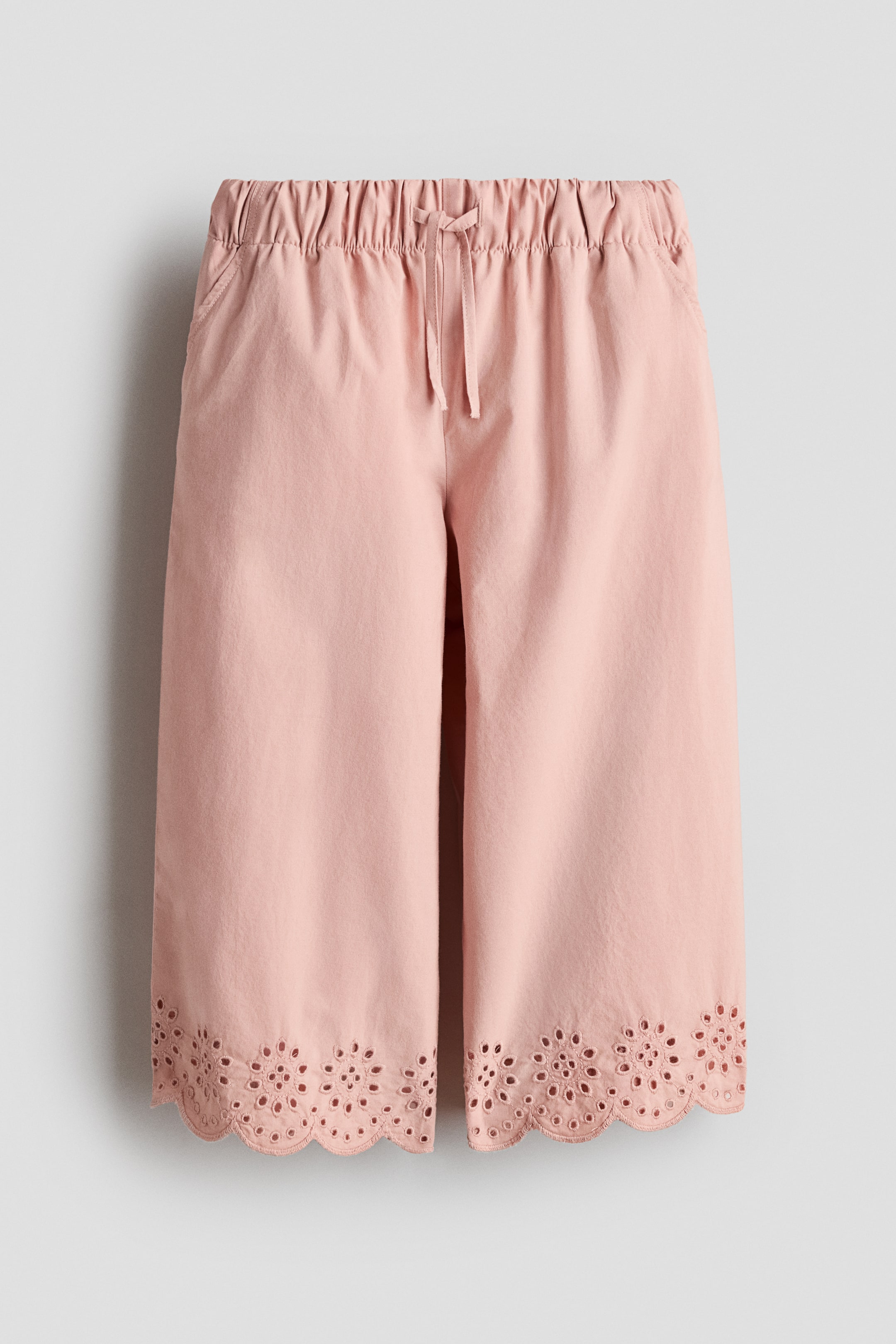 View larger image: Pink trousers, showcased in a FRONT view, crafted from soft cotton poplin. They boast an elasticated, drawstring waist, diagonal side pockets, and wide legs adorned with scalloped hems detailed with broderie anglaise.