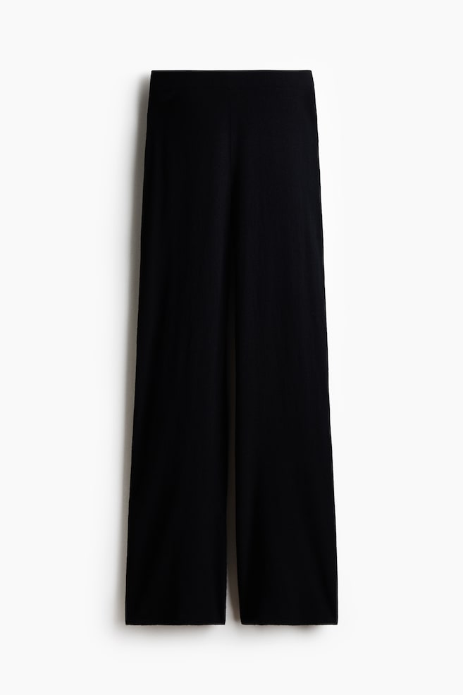 Black wide-leg knit trousers are displaying a clean-lined, high-rise elasticated waistband and a smooth, fluid texture. The trousers are falling straight down in a relaxed, wide-leg silhouette. - 1