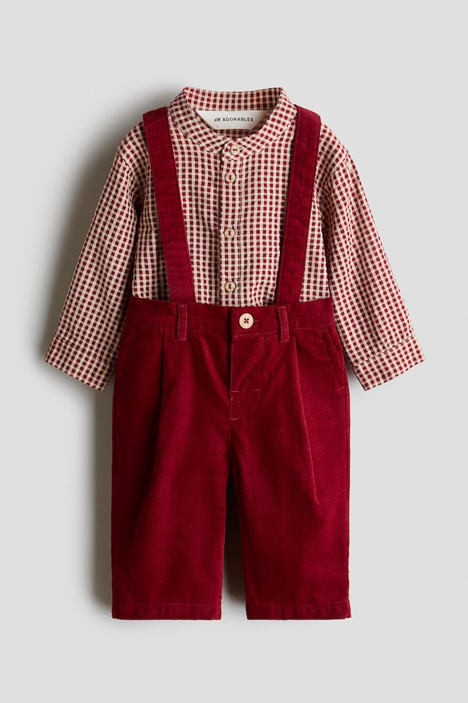 This front-facing view displays a children's outfit consisting of a red and white gingham-patterned long-sleeved shirt with a band collar. The shirt is paired with deep red corduroy trousers featuring matching suspender straps and a single-button fastening at the waist. - 1