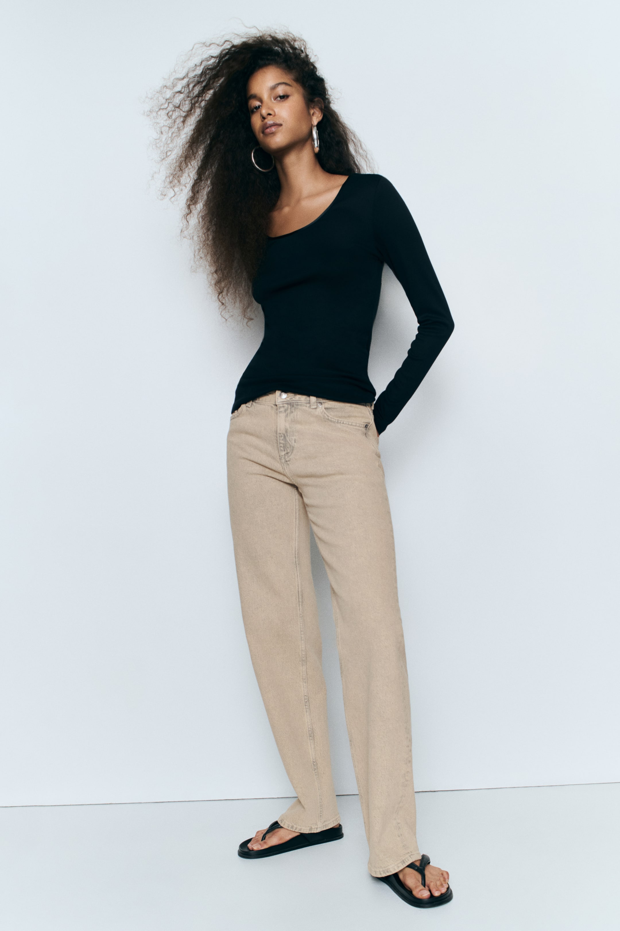 View larger image: The model is wearing beige five-pocket denim jeans with a regular waist, a slim fit through the hips, and a looser straight-leg cut that falls to the feet. The jeans are styled with a black scoop-neck long-sleeve top and black flat sandals.