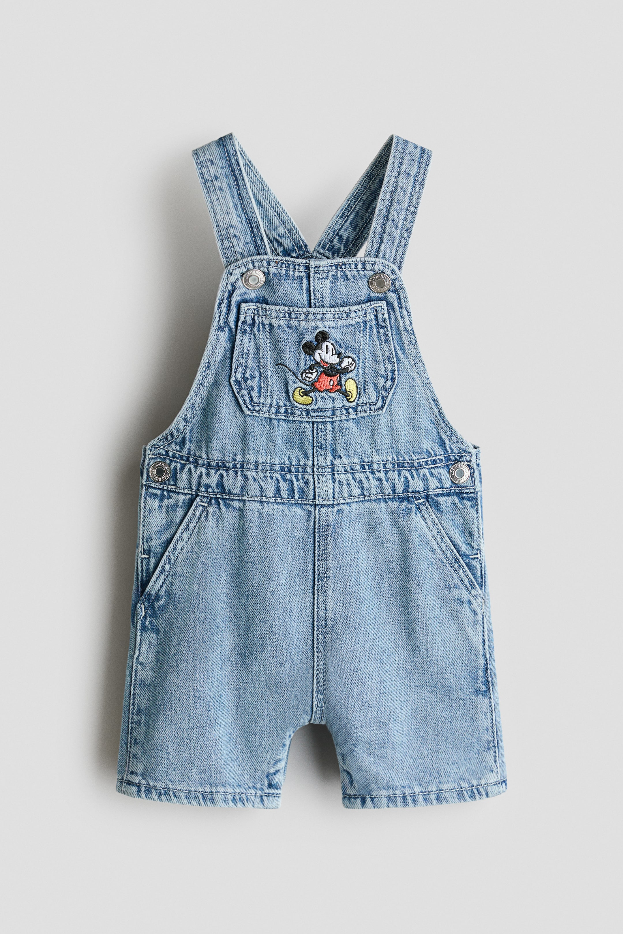 View larger image: Light blue denim overalls are showcasing at the front, featuring adjustable shoulder straps with metal buttons. The bib is highlighting a patch pocket with an embroidered Mickey Mouse, while the shorts include two side pockets and stitched hems.