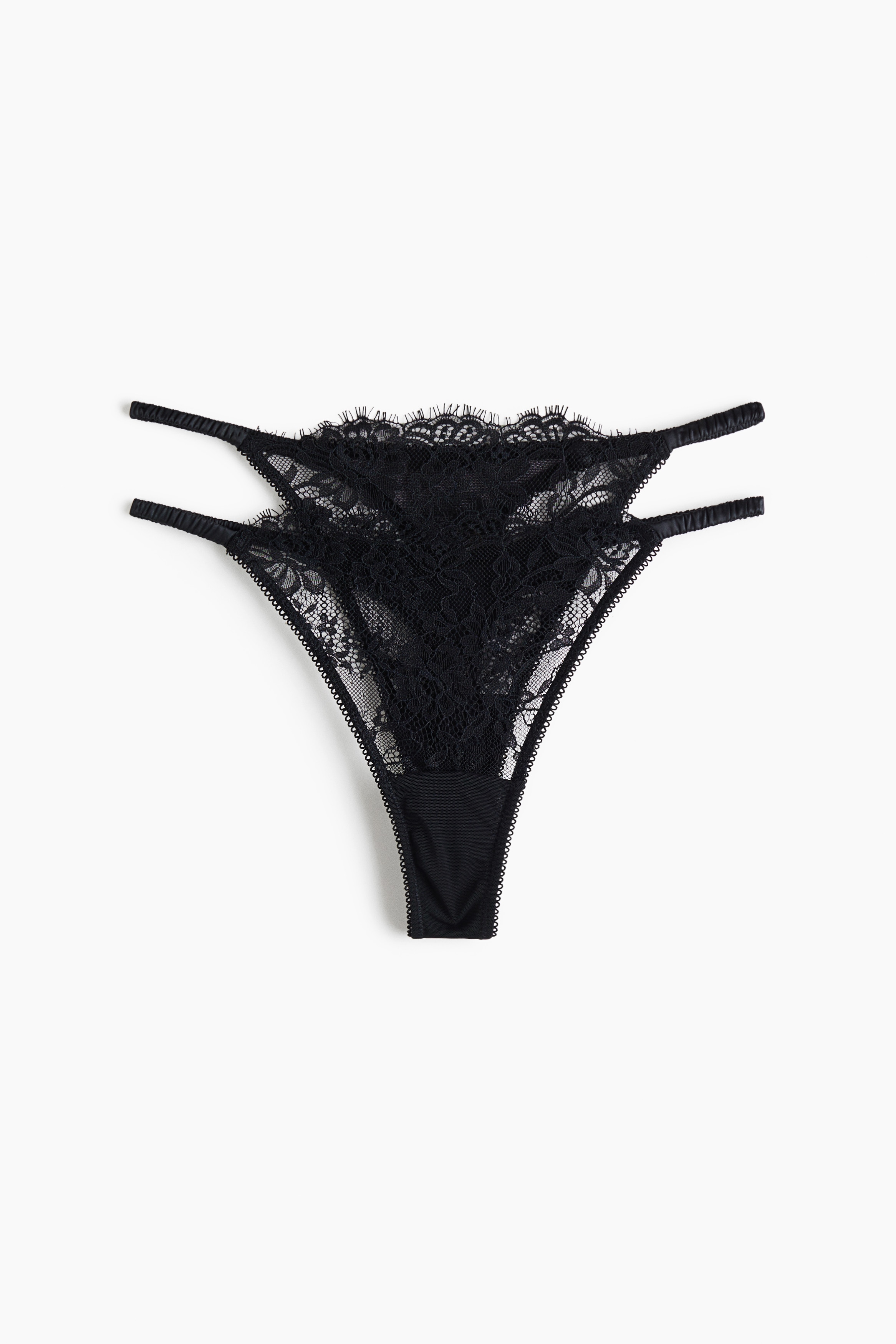 A black lace G-string featuring a low-rise waist and narrow side straps is worn by the model, paired with a matching black lace bralette. The model is posing in a seated, crouched position, showcasing the fit and details of the lingerie set.