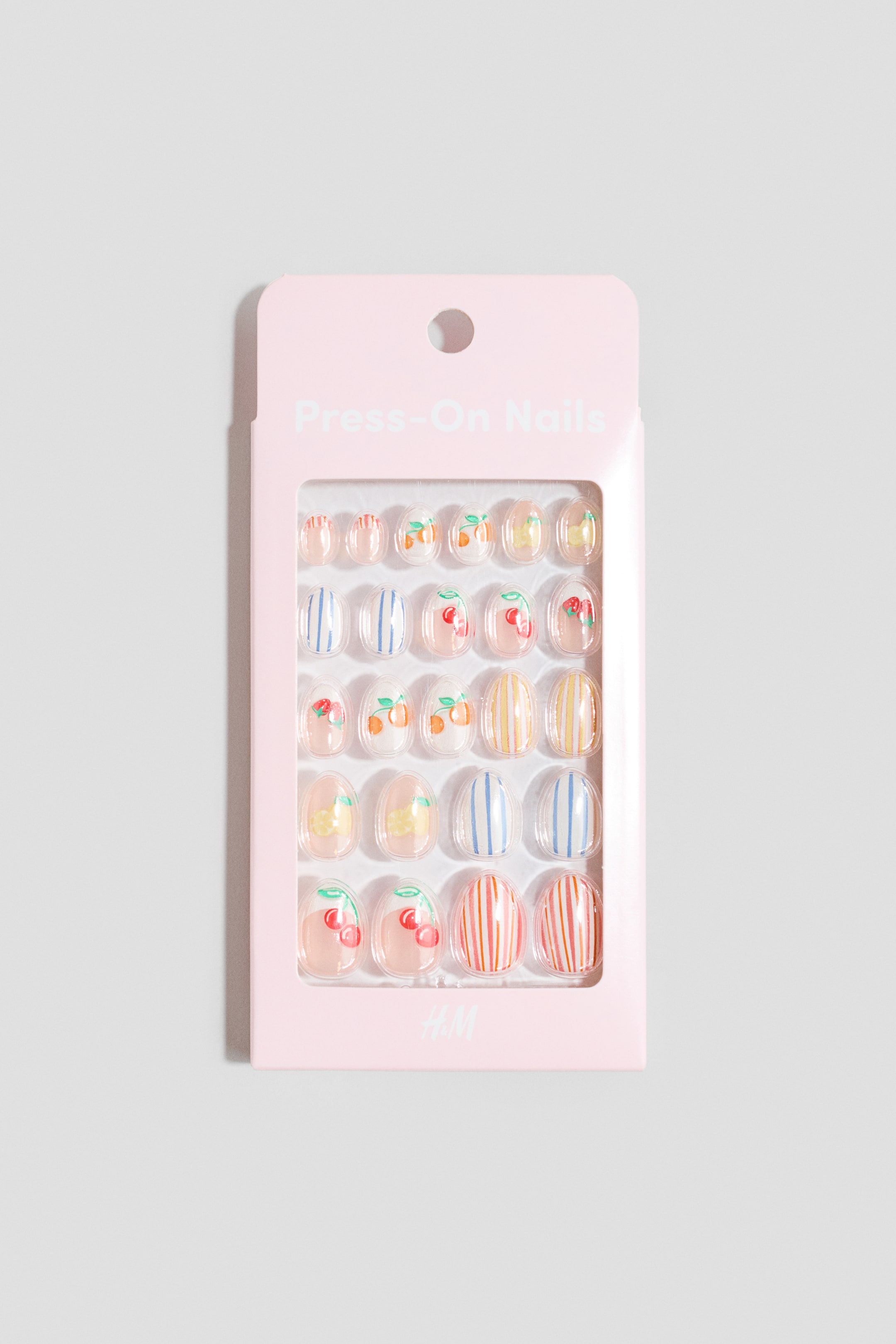 Agrandir l'image: Les ongles à presser sont présentés dans un emballage de vente au détail rose, avec une fenêtre transparente et les mots "Press-On Nails" imprimés en haut et "H&M" en bas. À l'intérieur, plusieurs ongles transparents à presser mettent en valeur divers motifs, notamment des cerises, des oranges, des rayures bleues, jaunes et rouges.