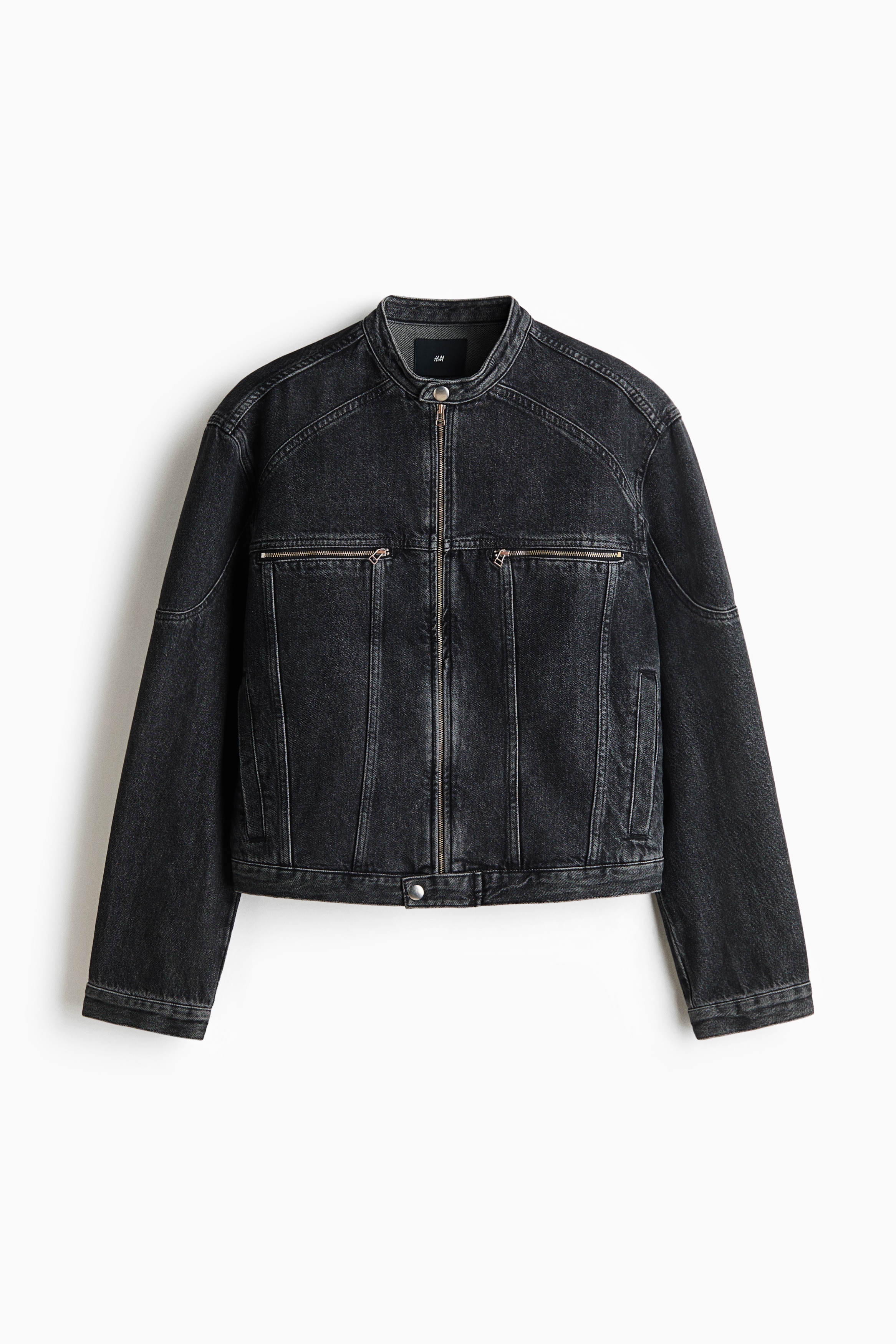 La modelo lleva una chaqueta corta de denim negro rÃ­gido de algodÃ³n, con cuello mao, cierre frontal de cremallera y bolsillos en el pecho con cremallera. La chaqueta se combina con una camiseta blanca y unos vaqueros negros a juego, mientras la modelo posa con una mano apoyada sobre una gorra.