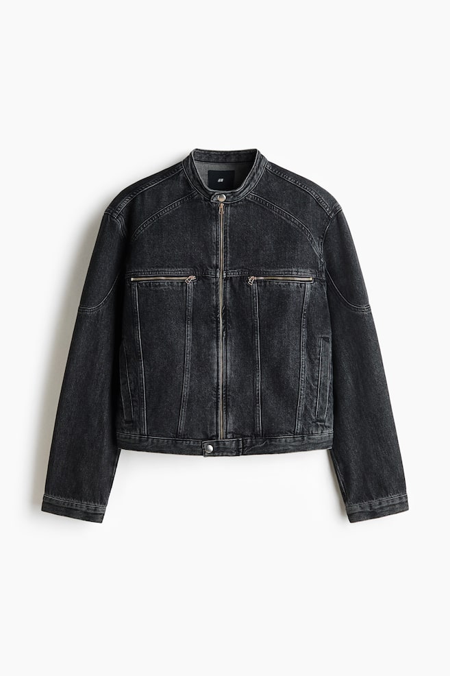 Eine kurze, schwarz gewaschene Denimjacke ist in frontaler Ansicht zu sehen und verfÃ¼gt Ã¼ber einen Stehkragen mit Druckknopf sowie einen durchgehenden FrontreiÃverschluss. Das Design zeigt markante TeilungsnÃ¤hte, zwei horizontale Brusttaschen mit ReiÃverschluss und vertikale Paspeltaschen an den Seiten. - 1
