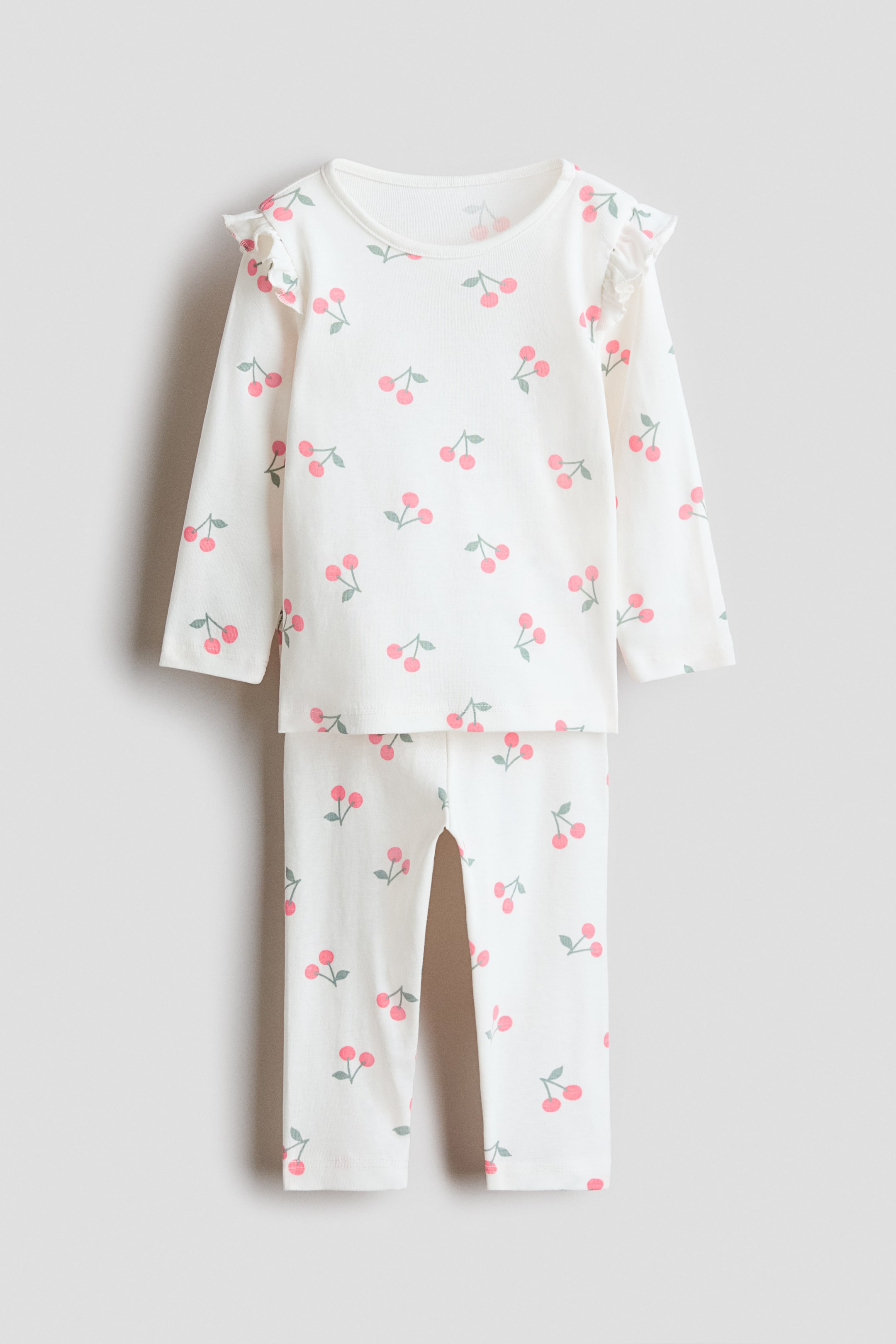 Προβολή μεγαλύτερης εικόνας: Ένα σετ πιτζάμες παρουσιάζει ένα τοπ με μακριά μανίκια και ταιριαστό παντελόνι πλήρους μήκους, και τα δύο σε εκρού βαμβακερό τζέρσεϊ με διάσπαρτο ροζ μοτίβο κερασιών, απεικονιζόμενα από μπροστά. Το τοπ διαθέτει στρογγυλή λαιμόκοψη και ραφές ώμων με βολάν.