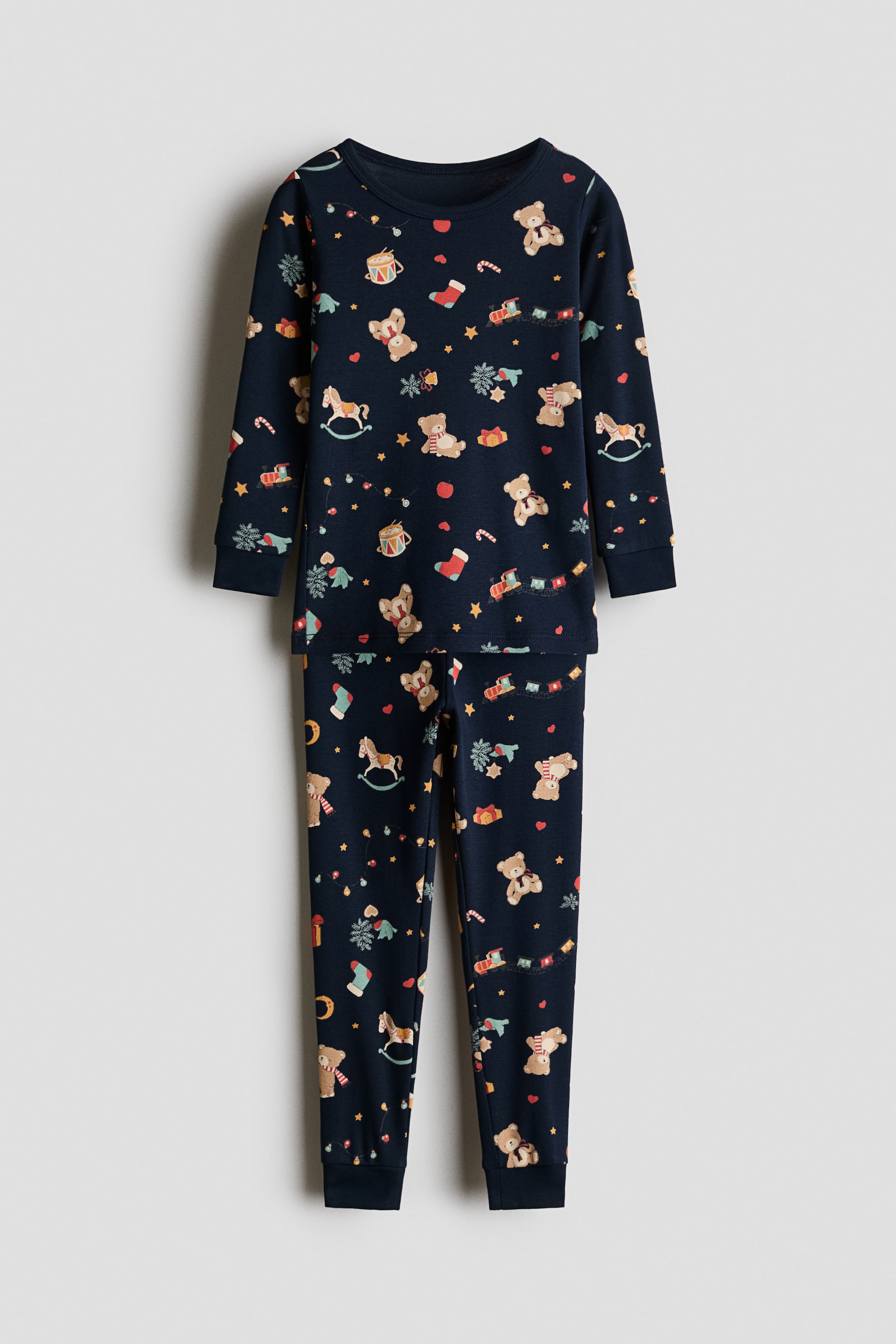 View larger image: A navy blue cotton jersey pajama set is shown from the front, featuring an all-over print of teddy bears, rocking horses, and festive motifs. The top features a round neckline and long sleeves with ribbed cuffs, while the matching pants feature ribbed cuffs at the ankles.