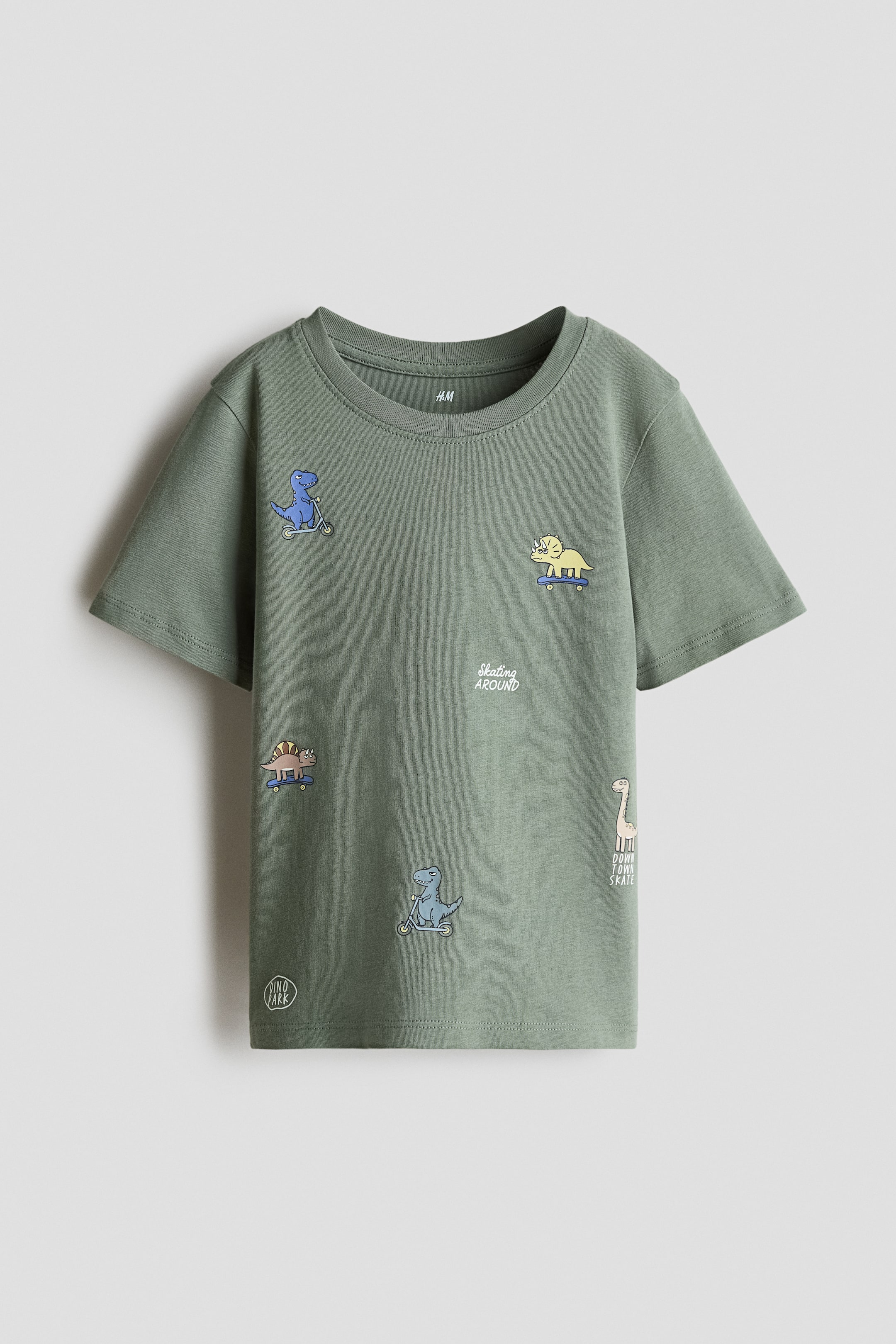 View larger image: A green T-shirt is laid flat, facing forward, featuring a ribbed crew neck and short sleeves. It showcases various cartoon dinosaur graphics riding scooters and skateboards, along with text phrases like "skating around" and "dino park."