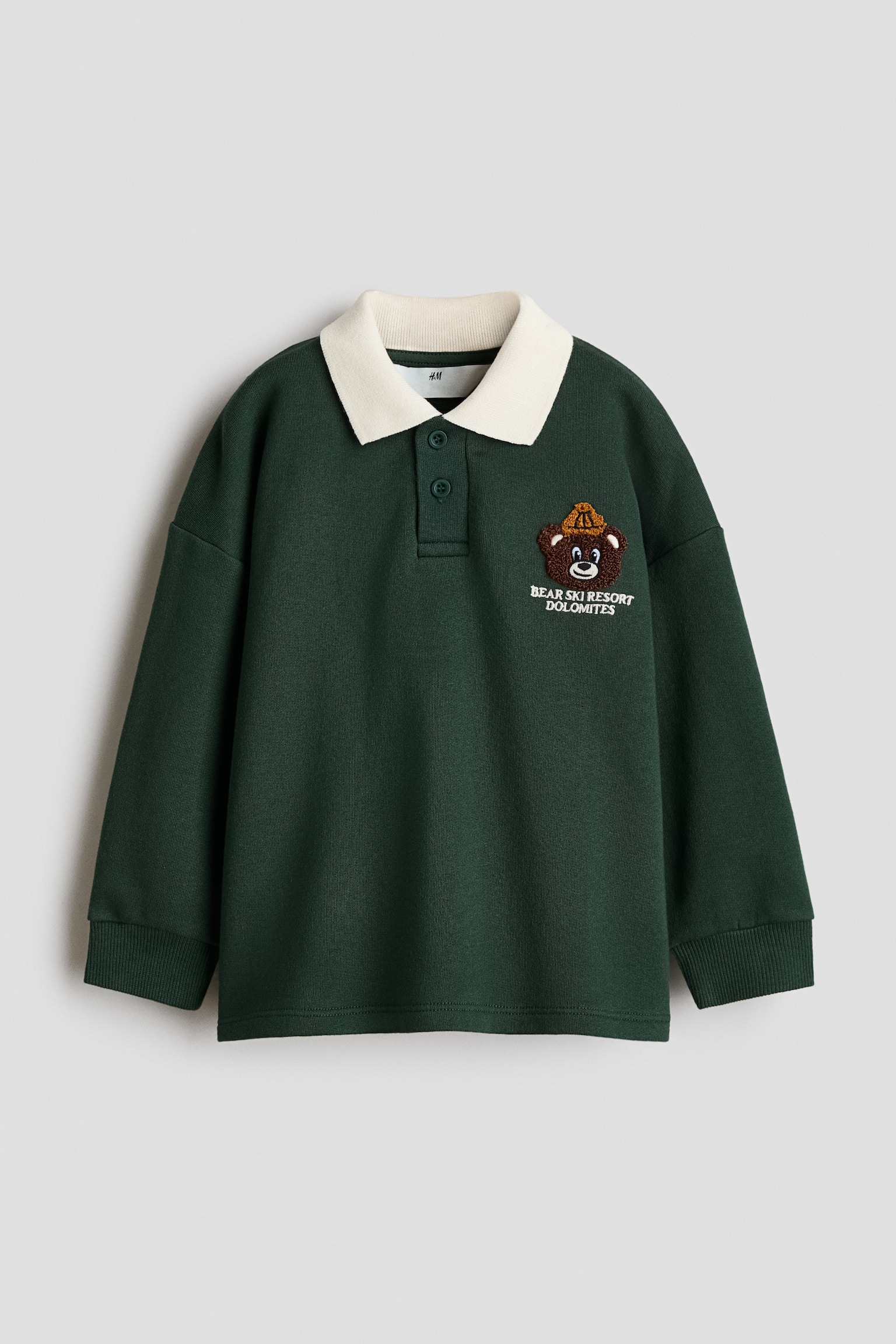 Vista frontal de una sudadera tipo polo de manga larga en verde oscuro, con cuello acanalado en color crema en contraste, tapeta de dos botones y hombros caÃ­dos con puÃ±os acanalados. Un parche bordado que representa un oso de dibujos animados marrÃ³n con un gorro de punto amarillo estÃ¡ colocado en el pecho izquierdo, encima del texto "BEAR SKI RESORT DOLOMITES".
