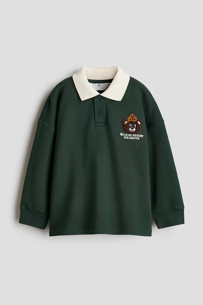 En framifrÃ¥n av en mÃ¶rkgrÃ¶n lÃ¥ngÃ¤rmad polotrÃ¶ja i sweatshirtmodell med kontrasterande ribbad krage i krÃ¤mfÃ¤rg, knappslÃ¥ med tvÃ¥ knappar och nedsÃ¤nkta axlar med ribbade muddar. En broderad patch med en brun tecknad bjÃ¶rn som bÃ¤r en gul stickad mÃ¶ssa Ã¤r placerad pÃ¥ vÃ¤nster brÃ¶st ovanfÃ¶r texten "BEAR SKI RESORT DOLOMITES". - 1
