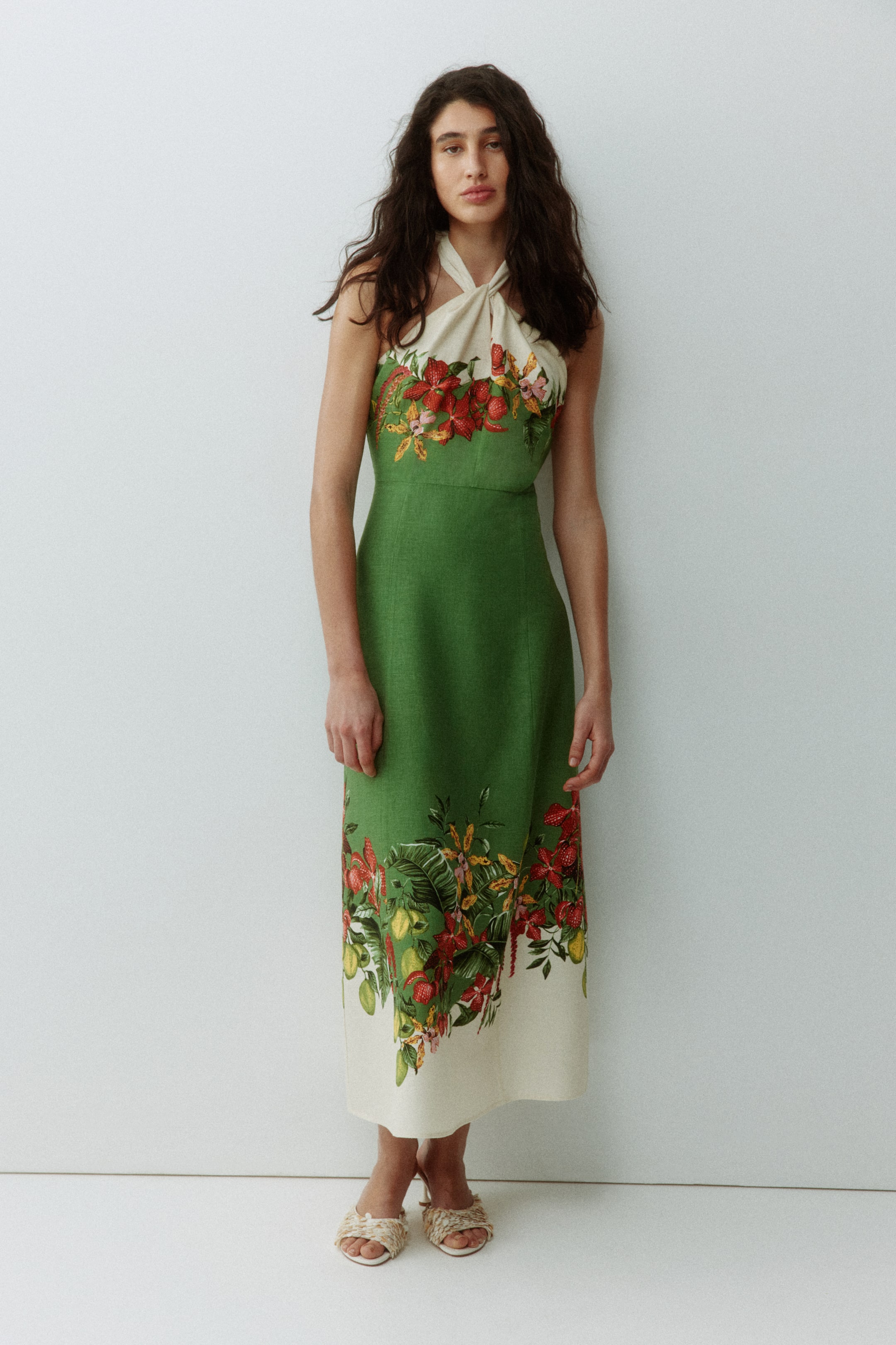 Agrandir l'image: La mannequin porte une robe mi-mollet en tissu tissé. La robe présente un corps vert avec une bordure crème à l'ourlet, un corsage ajusté et un col licou torsadé crème, avec un motif imprimé de fleurs rouges, de feuilles vertes et de fruits jaunes visible au niveau de l'encolure et de l'ourlet.