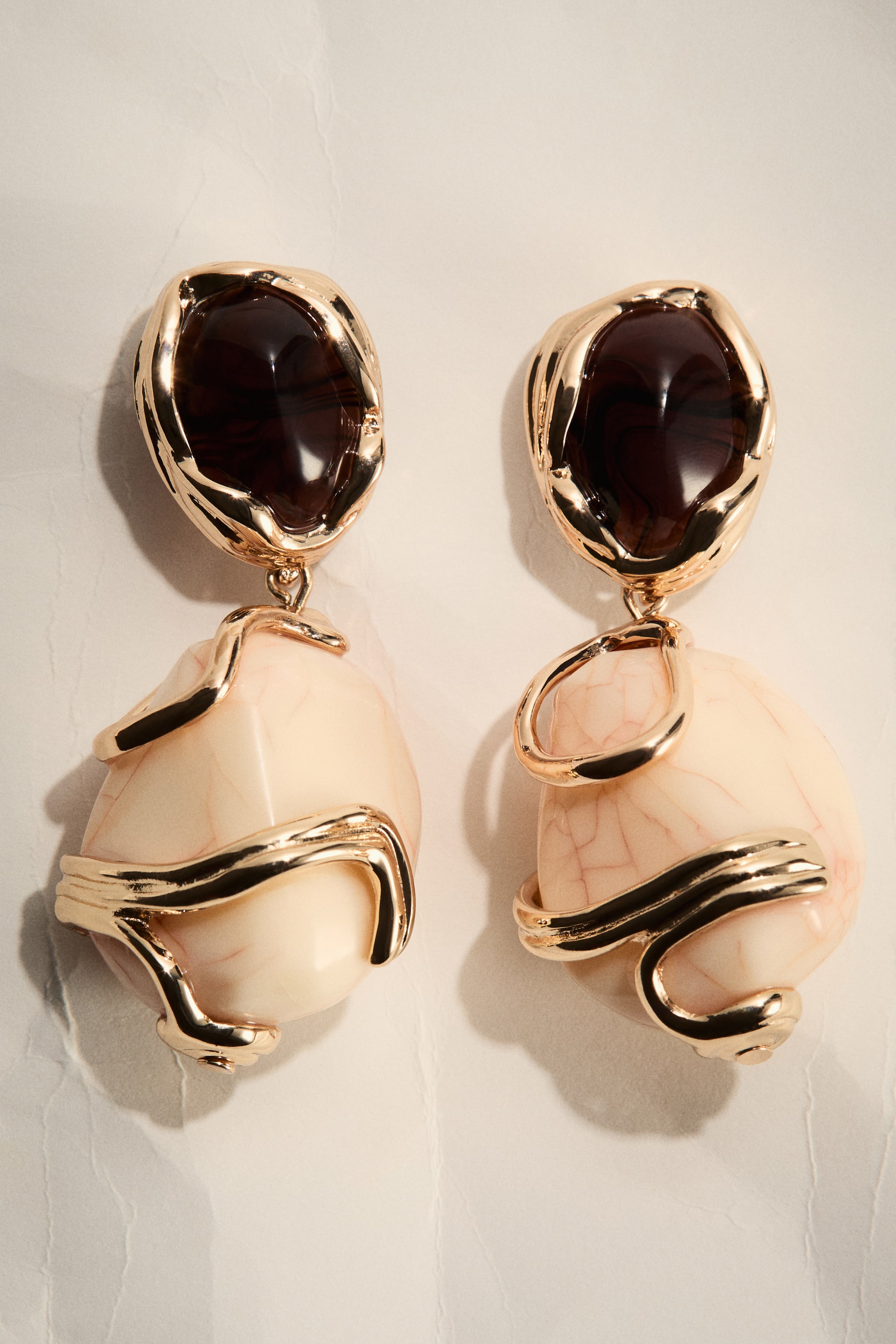 Nagyobb kép megtekintése: Egy pár lógó fülbevaló elölnézetből látható, mindegyik fülbevaló egy szabálytalan sötétbarna áttetsző felső elemmel rendelkezik, amely egy texturált arany tónusú foglalatba van ágyazva, és egy nagyobb, szabálytalan világos bézs alsó kőhöz kapcsolódik. Ez az alsó kő vörösesbarna erezetet mutat, és részben egy szobrászati arany tónusú fém szalaggal van körbevonva.