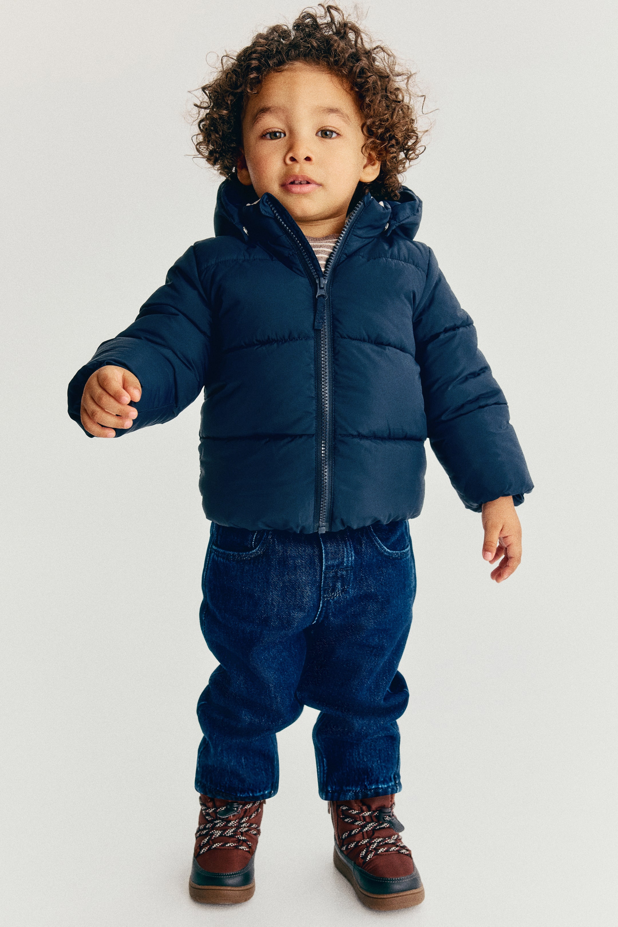 Größeres Bild ansehen: Eine marineblaue, gepolsterte Pufferjacke wird in Frontansicht gezeigt und verfügt über horizontale Steppnähte, einen Frontreißverschluss, einen hohen Stehkragen und eine Kapuze. Die Jacke ist an einem Kind mit dunkel gewaschener Jeans und braunen Winterstiefeln kombiniert.