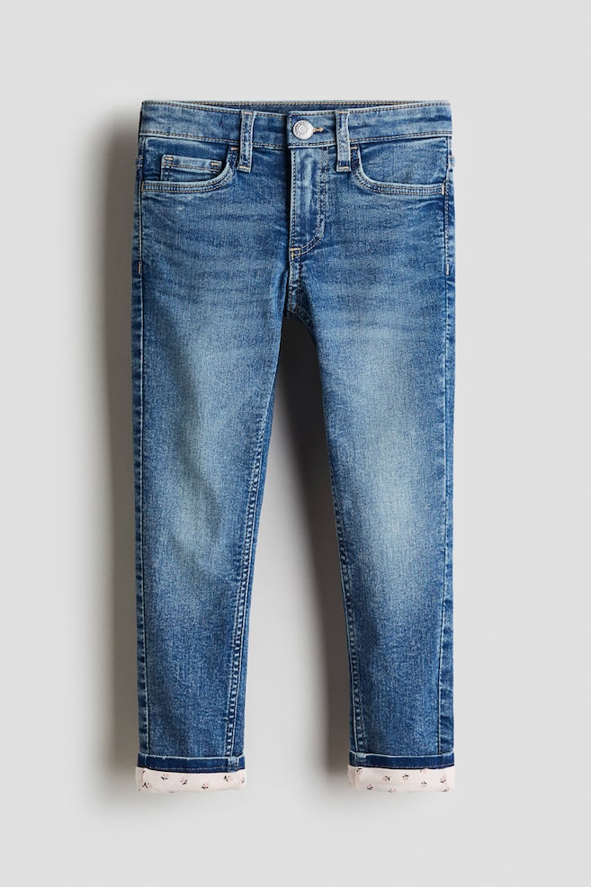 Ein Paar blaue Denim-Jeans wird von vorne präsentiert, mit einer engen Passform und einem 5-Taschen-Stil. Die Jeans zeigen eine mittlere Waschung mit subtilen Verblassungen, während die Beinabschlüsse hochgekrempelt sind und ein helles Futter mit einem kleinen Blumenmuster enthüllen. - 1