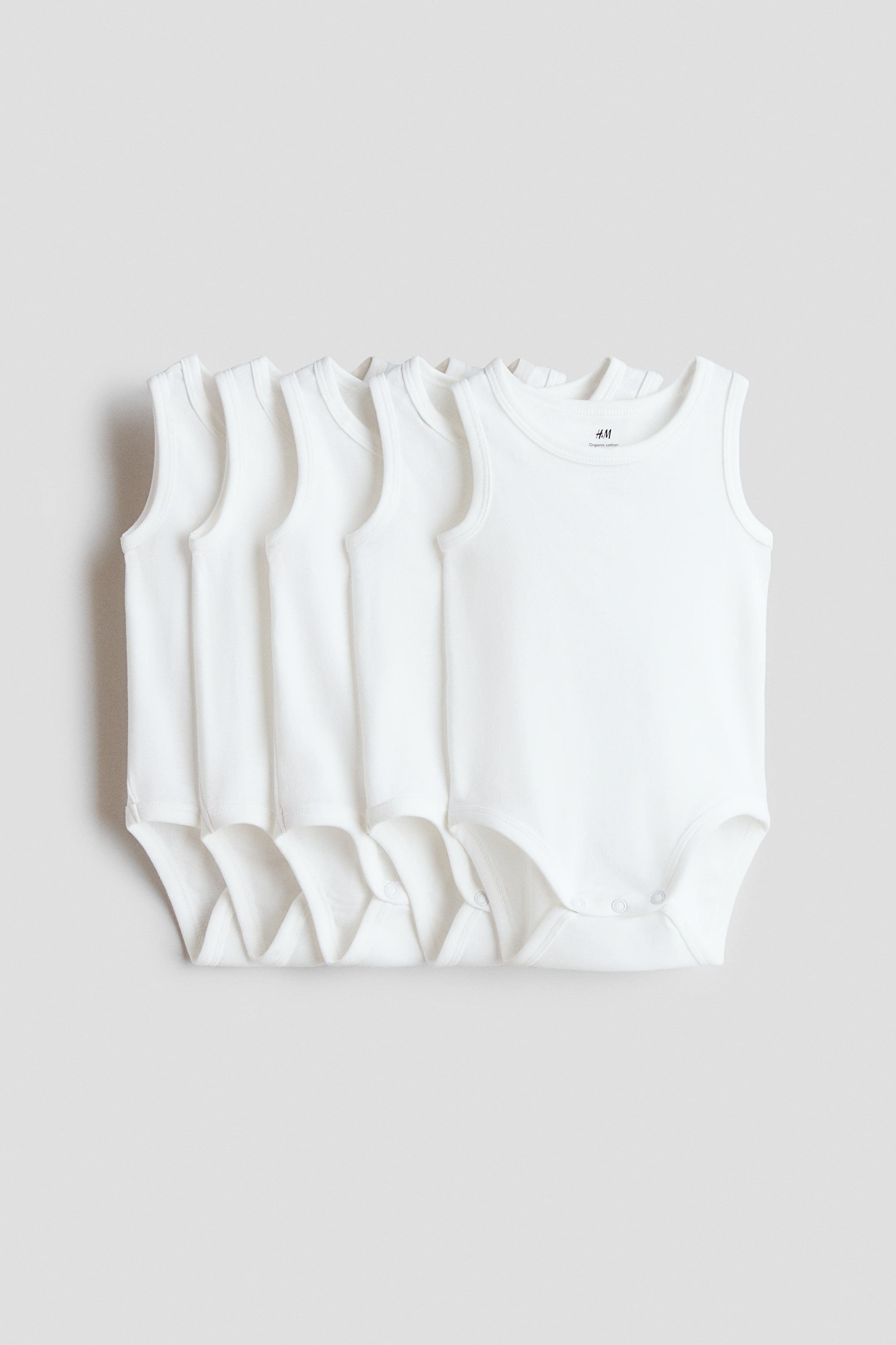 View larger image: "Sleeveless bodysuits in soft cotton jersey with press-studs at the crotch are displayed in a tidy line, with their white fronts and round necklines clearly facing forward."