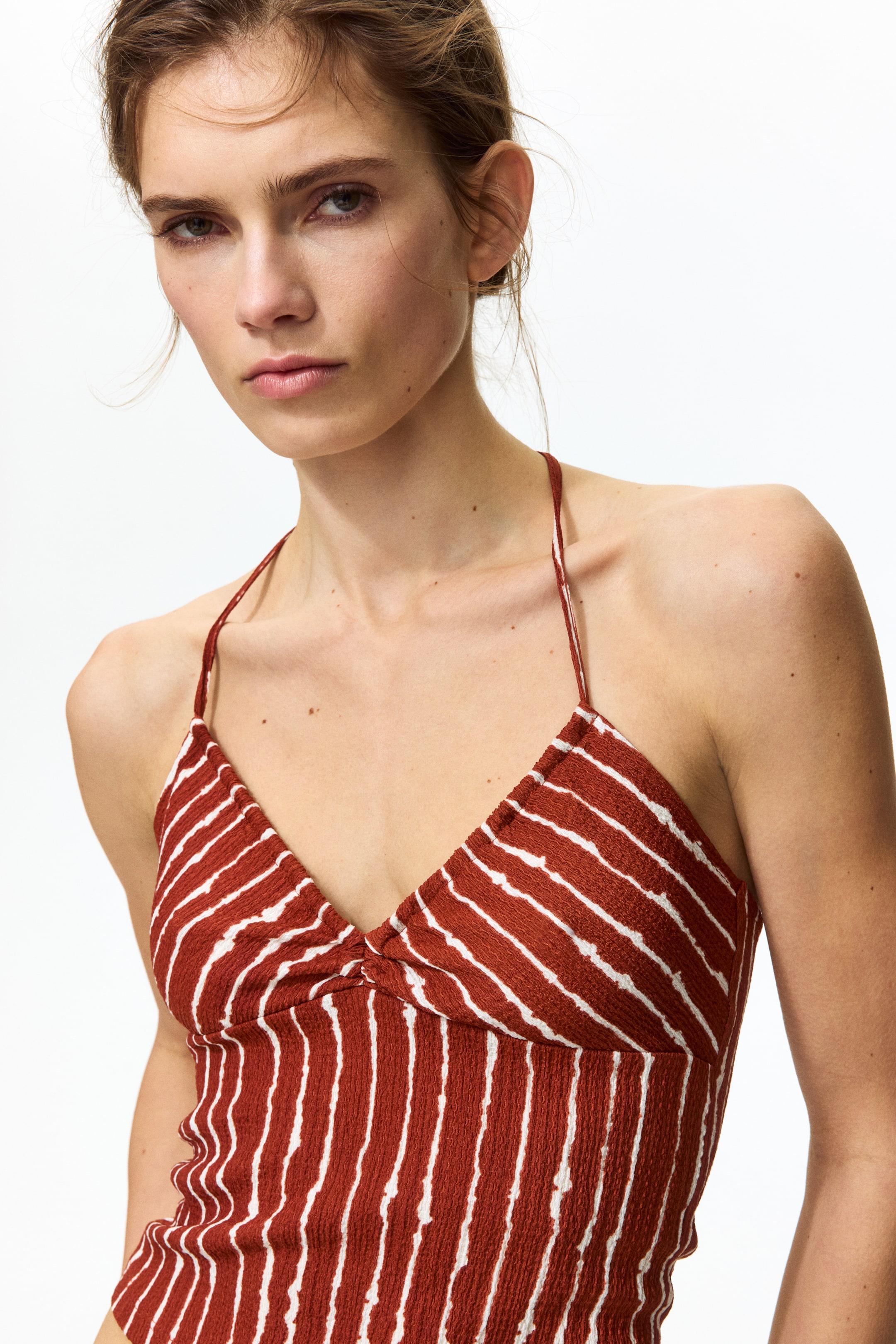Ampliar la imagen: Un top halter color óxido muestra rayas horizontales irregulares blancas sobre una tela texturizada, con un escote en V que presenta un detalle central fruncido y finos tirantes halter.