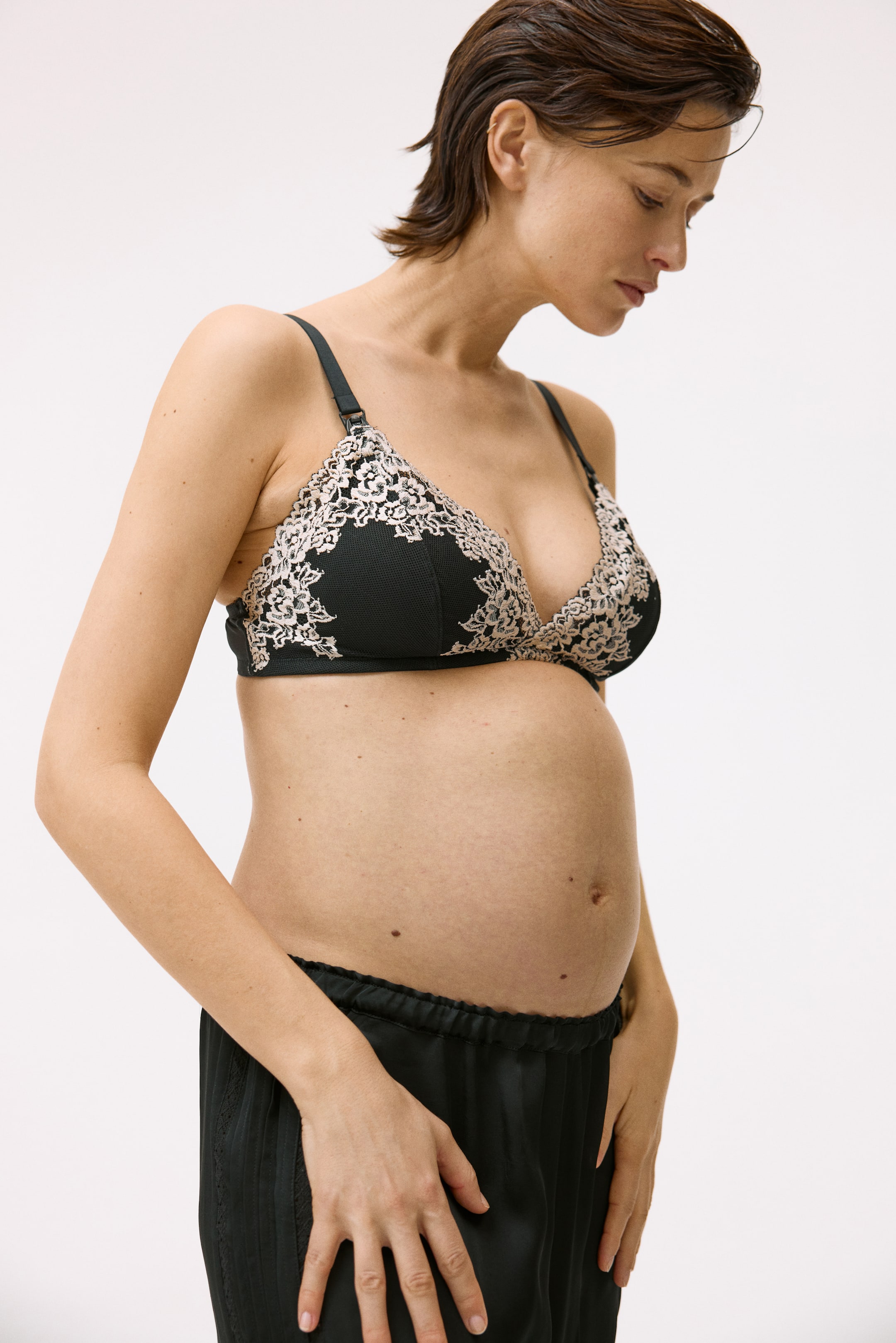 View larger image: The black bra showcases intricate pale lace embroidery adorning the cups and extending onto the band. It features black adjustable straps with a nursing clip visible on the left strap, and is presented from a slight front-right angle.