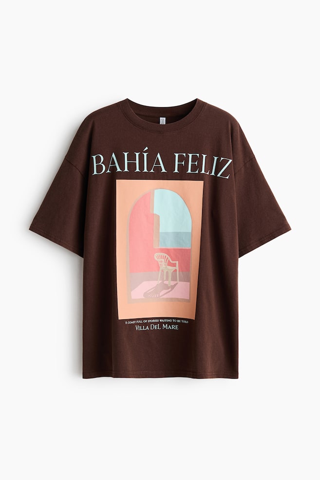 A dark brown oversized t-shirt is showcasing its front, featuring a ribbed crew neck and short sleeves. The front displays 'BAHÍA FELIZ' in light blue text above a rectangular graphic depicting an archway scene with a chair, peach and pink walls, a light blue opening, and small white text reading 'A COAST FULL OF STORIES WAITING TO BE TOLD VILLA DEL MARE' at the bottom. - 1