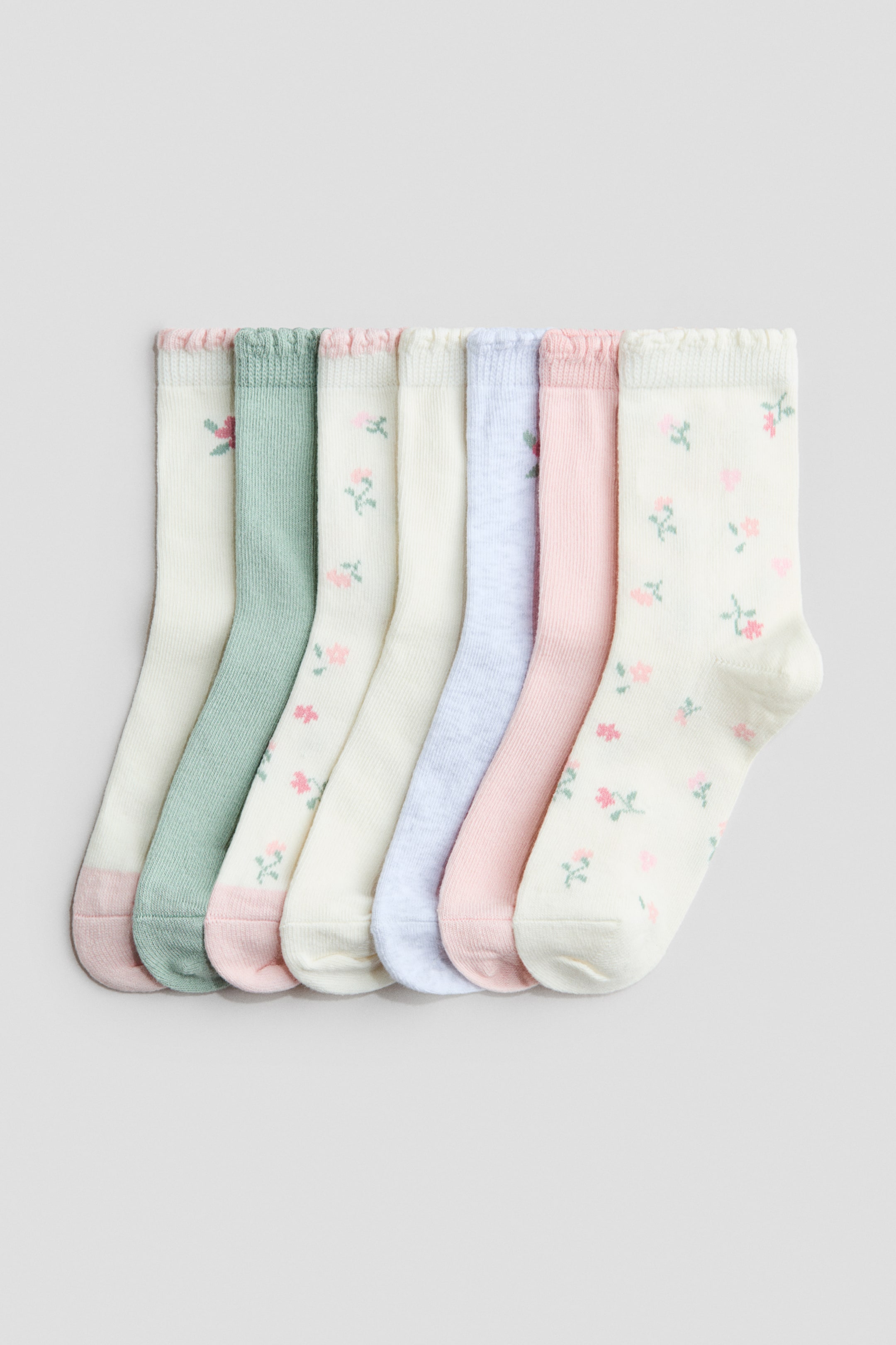 View larger image: Fine-knit socks in a soft cotton blend with elasticated tops, presented as a set of seven, arranged front-on and slightly overlapping horizontally. Three of the socks feature small pink and green floral patterns on a cream or light pink base, while four solid-coloured socks appear in light green, cream, light blue and light pink; all have contrasting-coloured heels and toes.