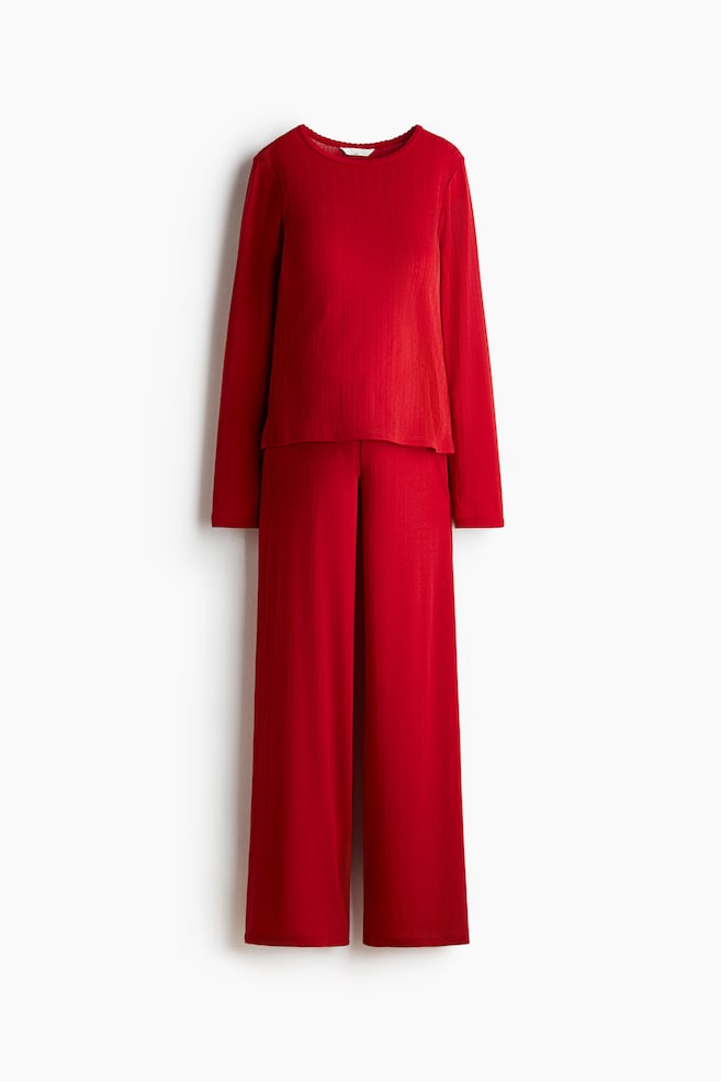 A red two-piece set is visible, featuring a long-sleeved top with a round neckline and a scalloped edge, paired with matching wide-leg pants showcasing a pleated front. Both pieces are displaying a finely ribbed knit texture. - 1