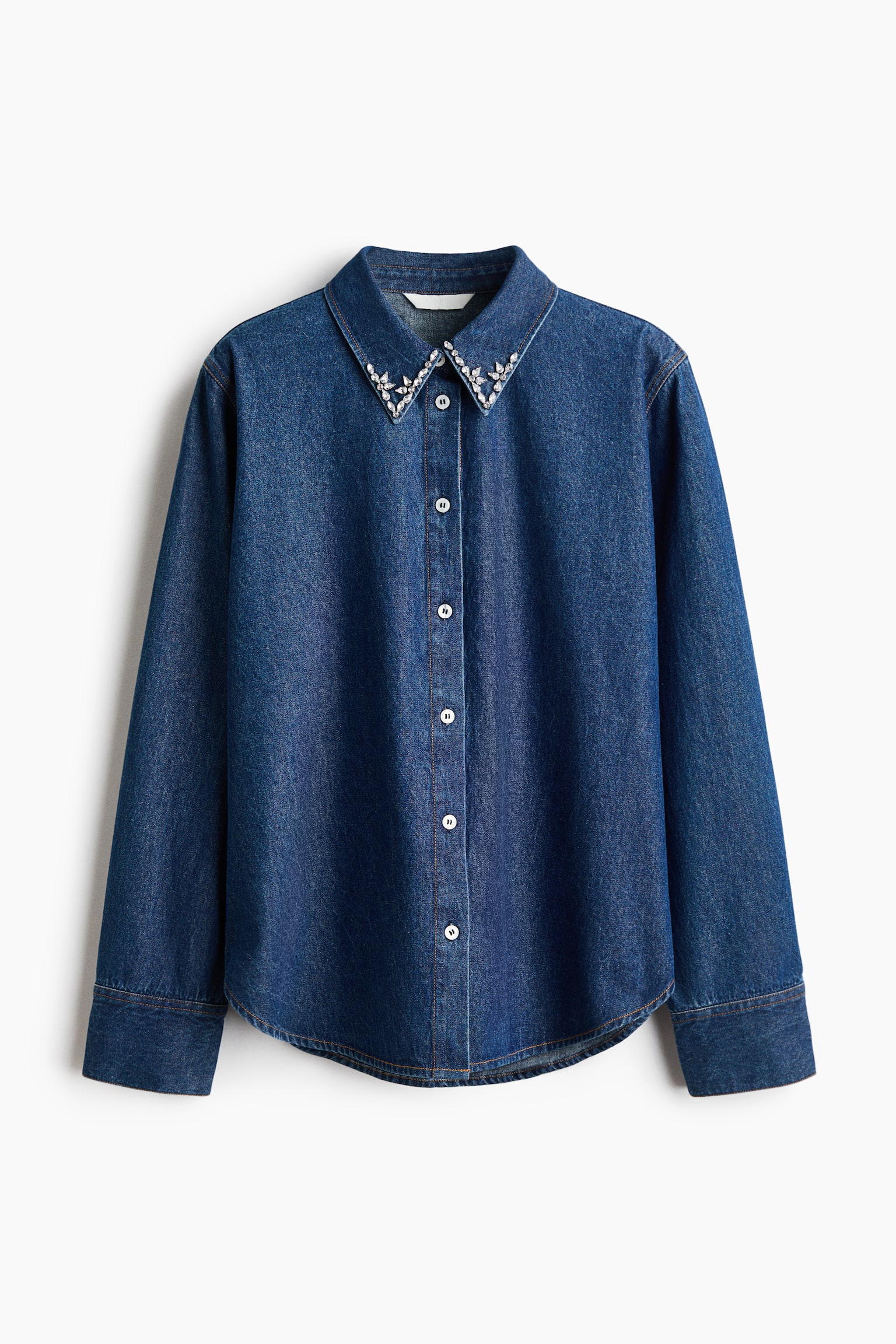 A long-sleeved blue denim shirt is shown from the front, featuring a pointed collar embellished with clear, sparkling rhinestones in a decorative pattern. The shirt has a front button-down closure with light-coloured buttons, long sleeves with wide cuffs, and a curved hem.