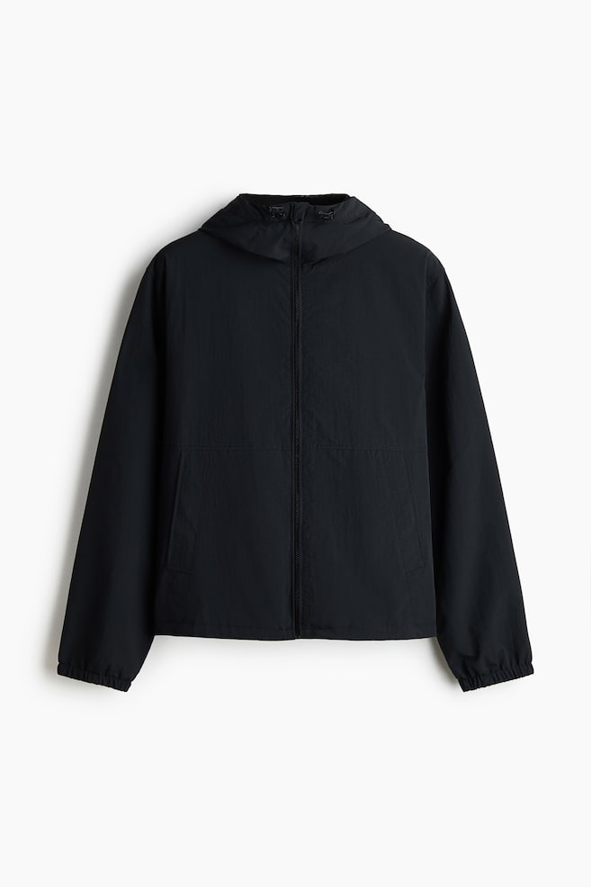 Está a ser apresentado um casaco corta-vento em nylon preto, FRENTE, com um capuz com cordão e travões, fecho de correr frontal de comprimento total, mangas compridas com punhos elásticos, e dois bolsos laterais integrados nas costuras frontais, terminado com uma bainha elástica. - 1